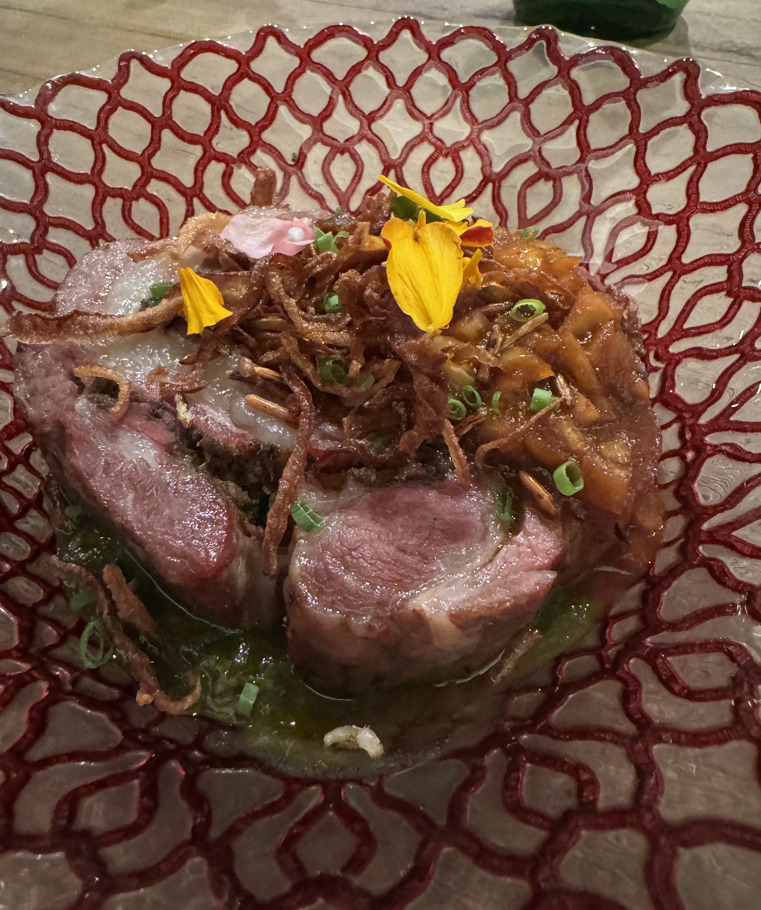 Very large slice of deboned lamb neck. It has segments of pink meat and white fat, swirled together. There is a green sauce beneath the lamb, and a golden-brown sauce with fried onions on top of it. There are yellow flower petals as a garnish.