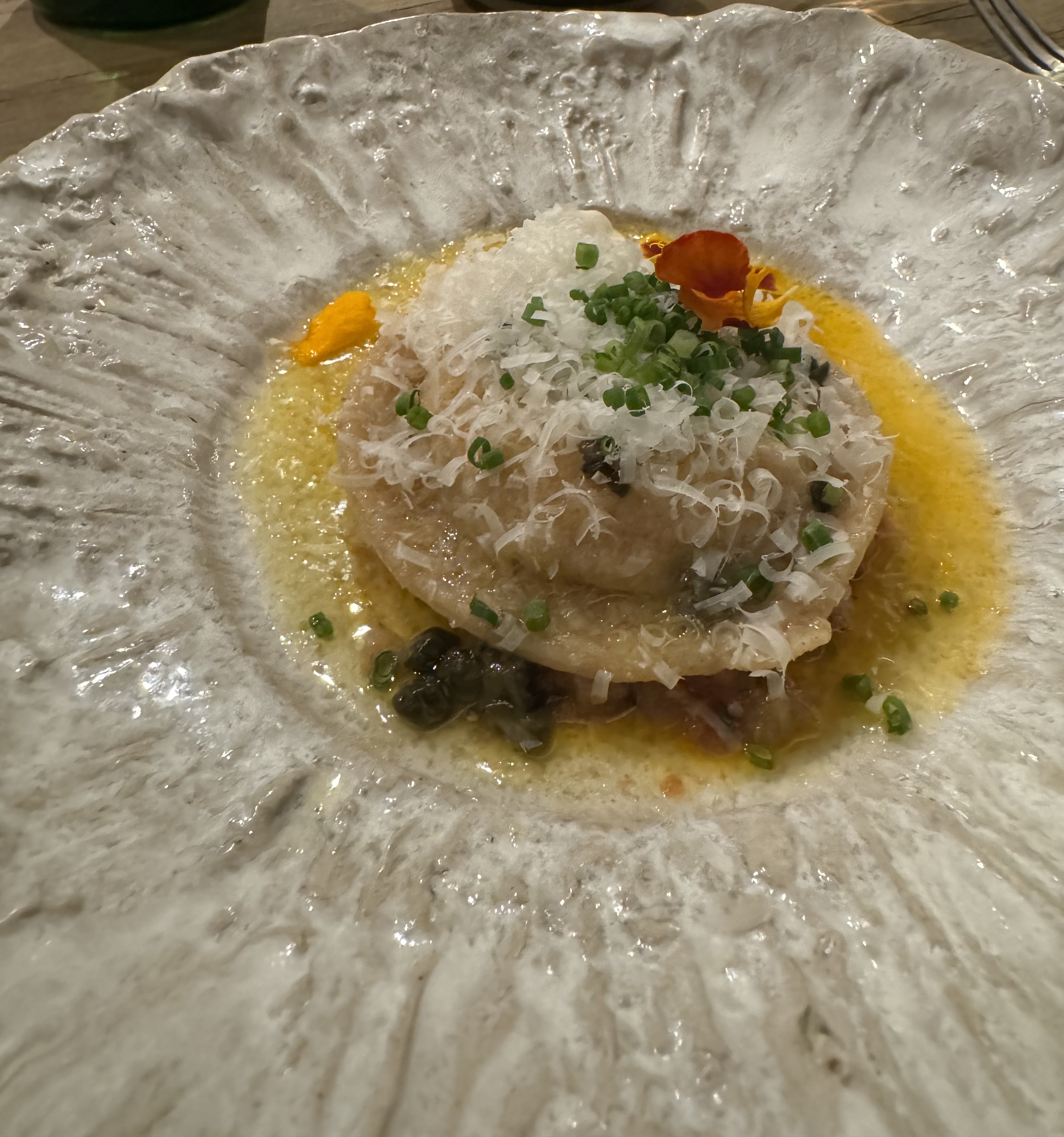 One single giant ravioli sitting in the middle of a large plate. There are capers below it, plenty of shredded cheese on top of it, and a deep yellow sauce around it. There are some orange flower petals and thin slices of chive as garnish.