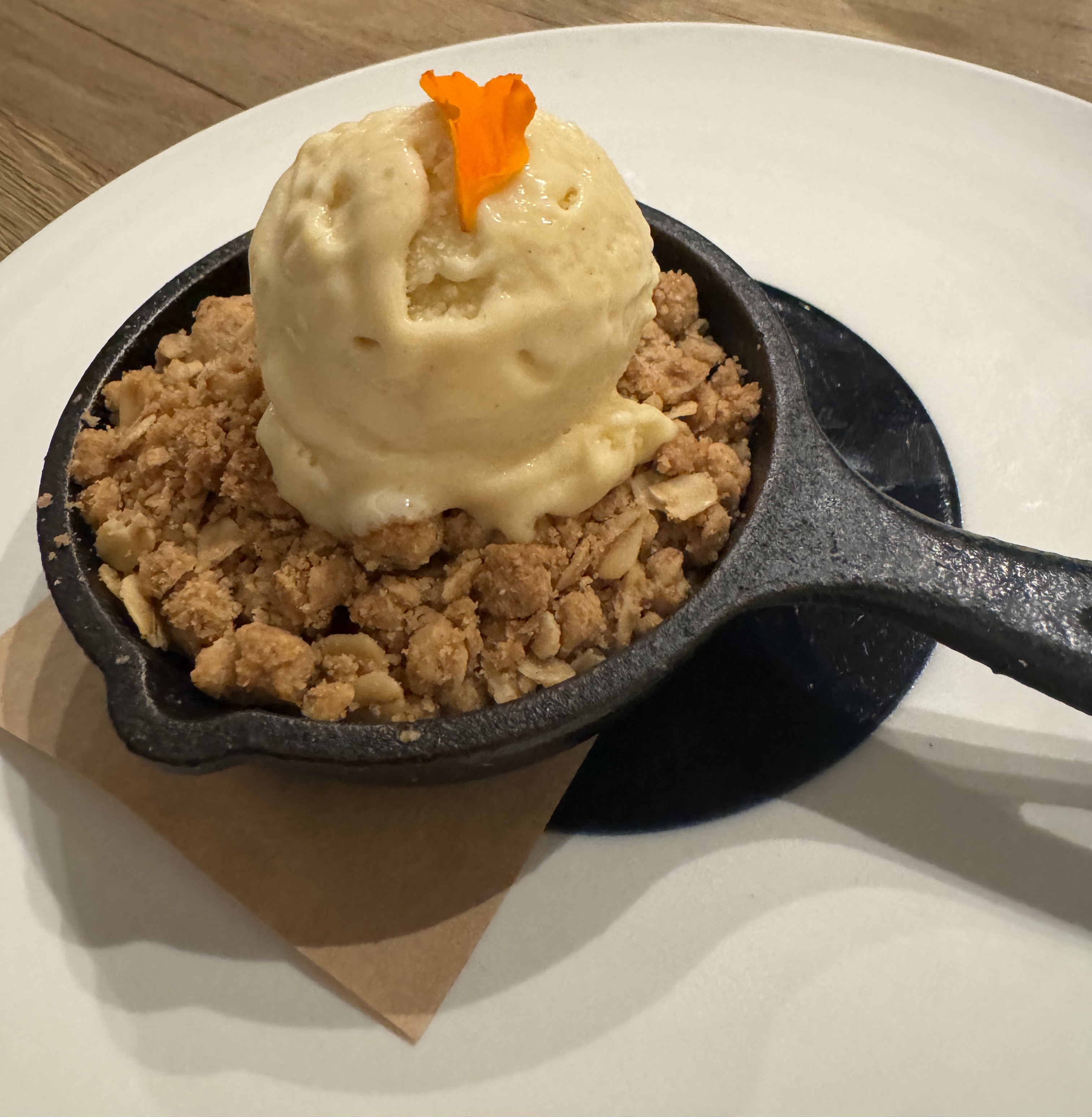 A tiny cast-iron skillet on a normal-size plate. It's filled with a crumble, and an off-white sort-of-yellow-ish scoop of ice cream is sitting on top. There is a single orange flower petal as garnish.