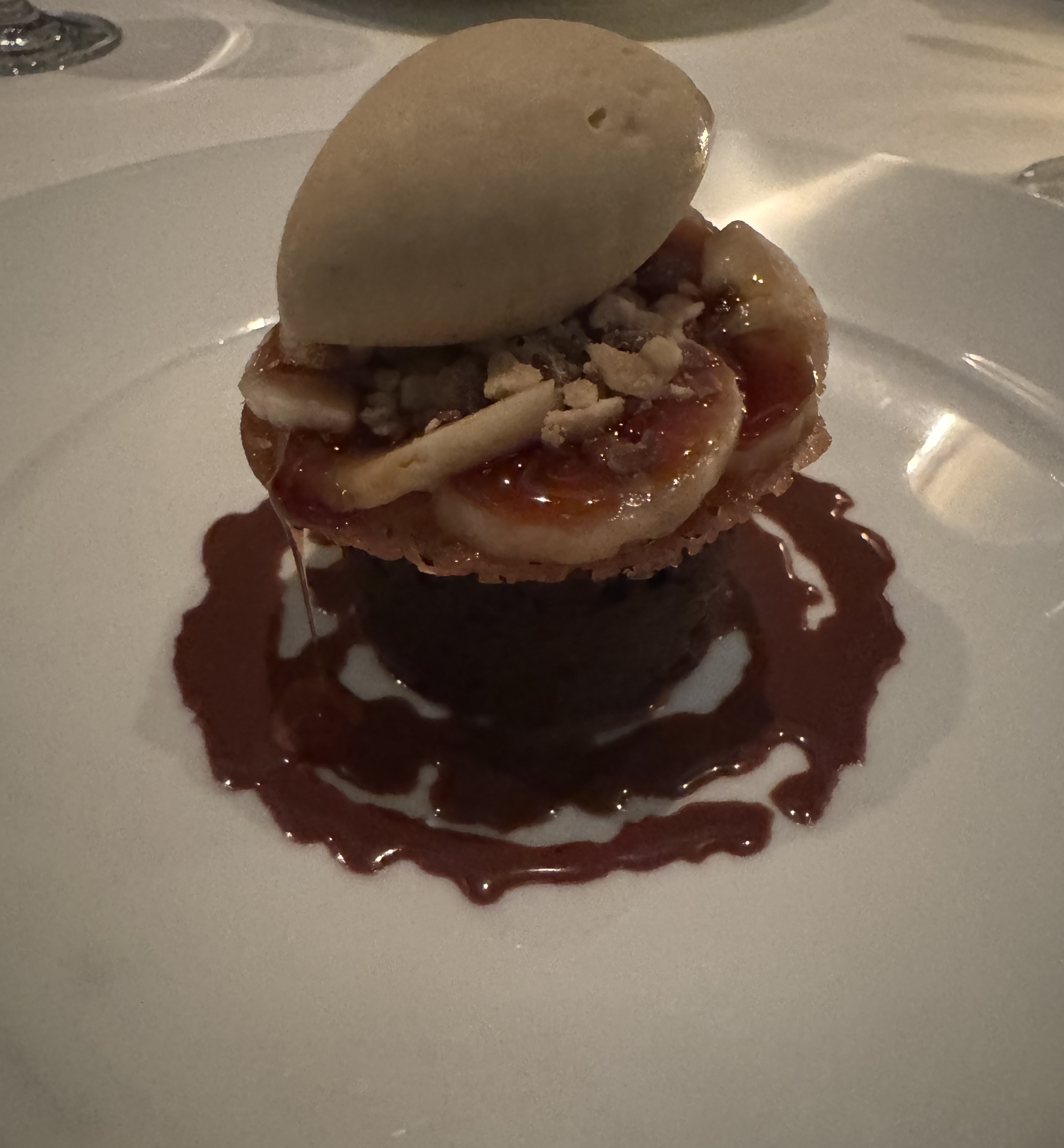 Stack of sweet treats. The bottm of the stack is a dark cylindrical cake, surrounded by squirts of chocolate sauce on the plate. On top of the cylinder is a disc of light-brown cookie. There are several banana slices topped with caramel and nuts on top fo that. There is a light-brown quenelle of ice cream on the very top.