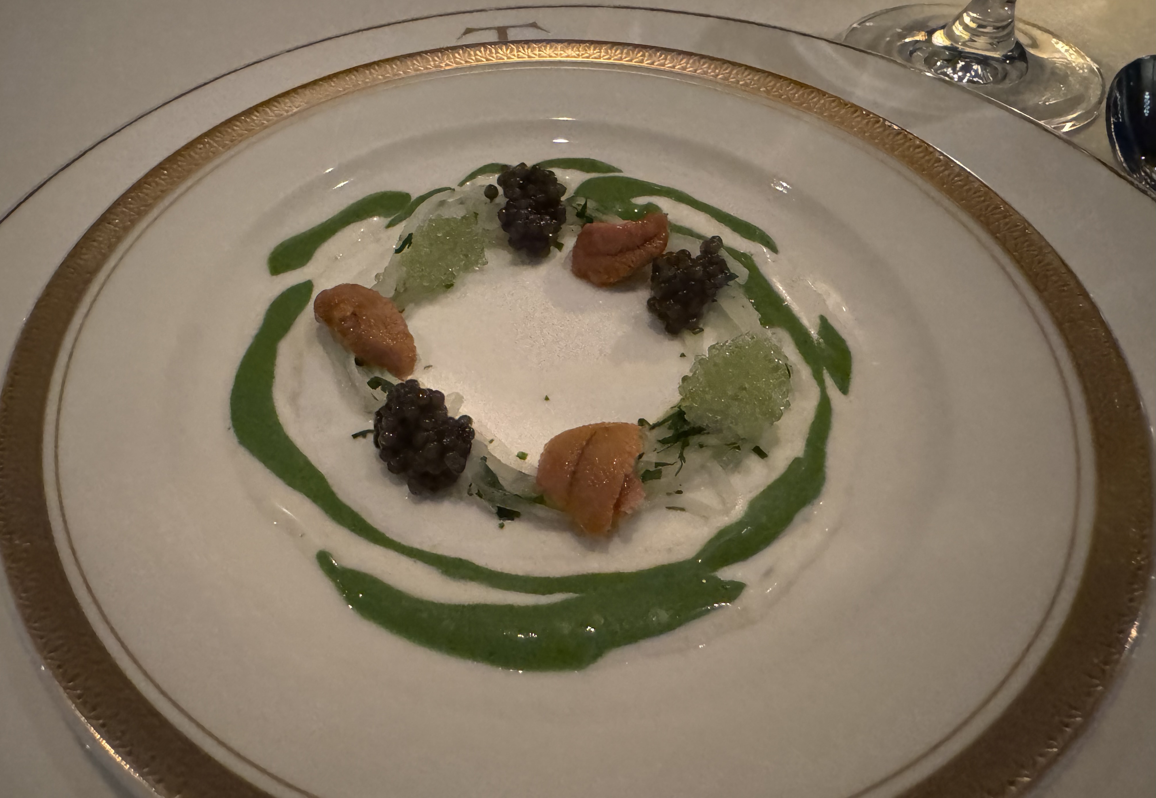 A smaller plate with a thick gold trim on top of the large Trotter's dinner plate. There is a ring of translucent shredded daikon radish with yellow tongues of uni, piles of caviar, and piles of finger lime. There's white sauce around the ring, with streaks of dark-green sauce.