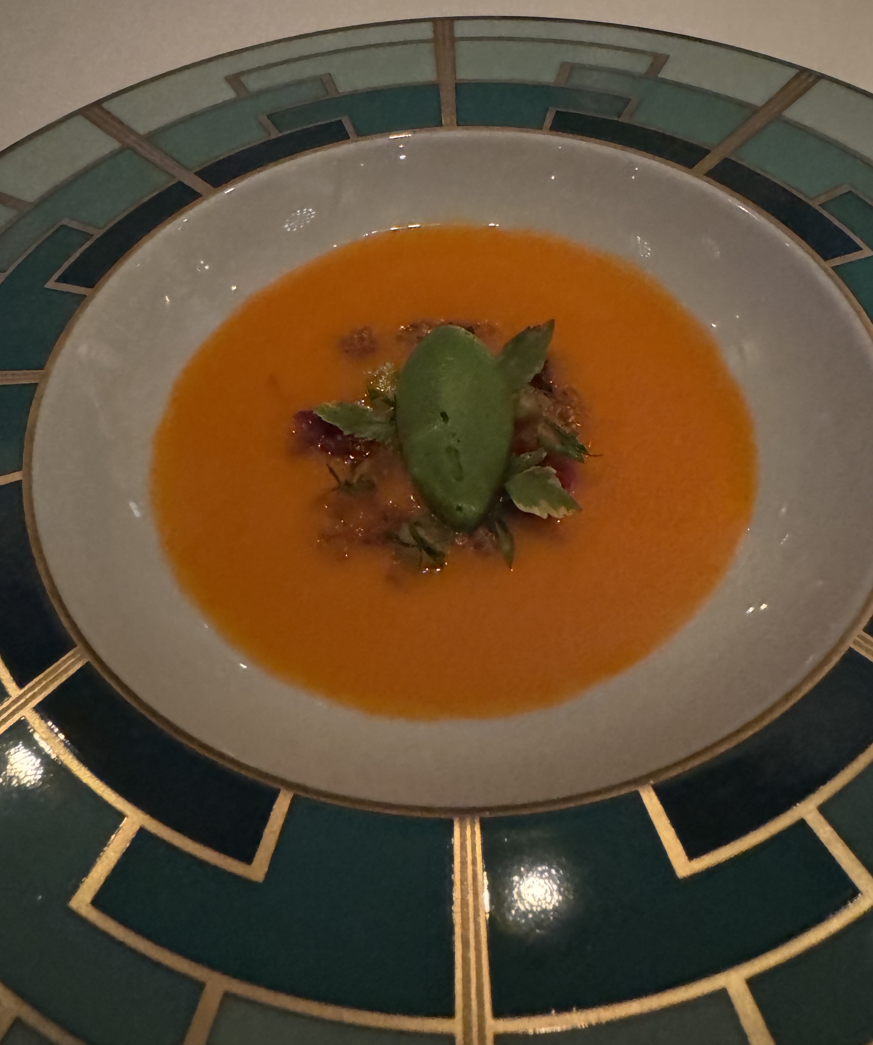 Shallow bowl with light-orange soup. In the center of the soup, there is dark-green quenelle of avocado & cilantro sorbet, several leaves, and some tiny chunks of bread.