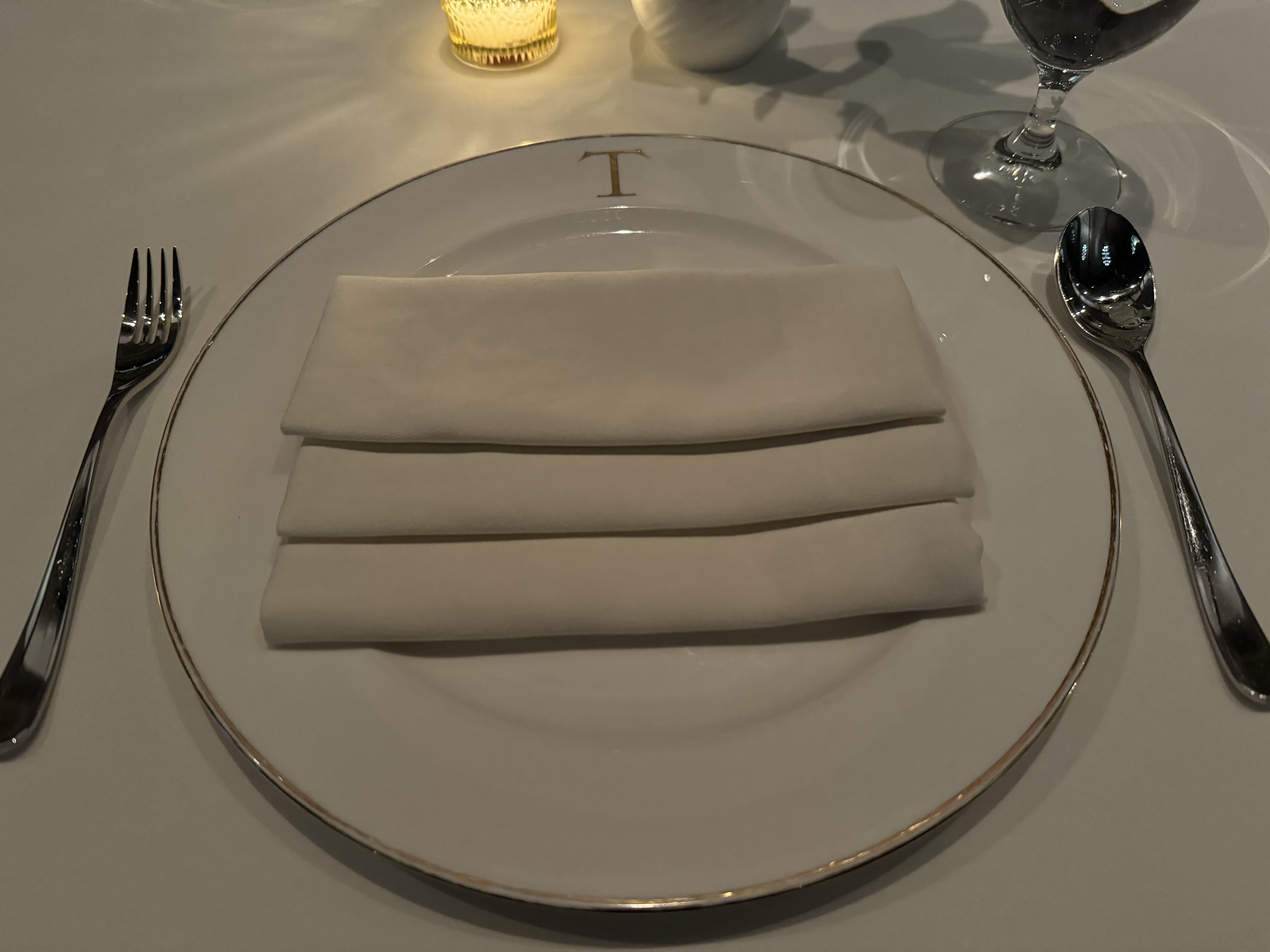 A table setting. There is a silver spoon & fork around a round plate. The plate is white, rimmed with gold trim. It has a golden 'T' at the top. There is a beautifully-folded napkin with three segments on the plate.