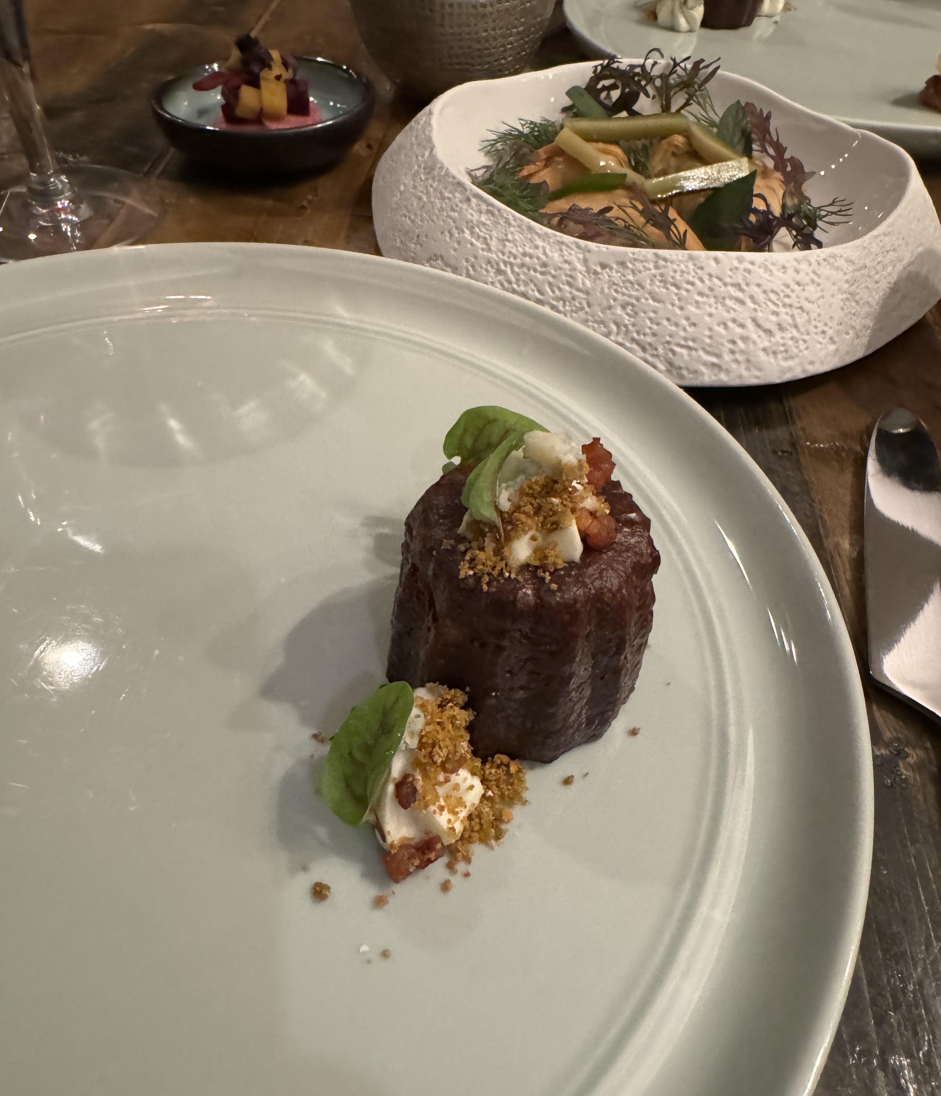 A huge plate with one small item on it: a canelé, with a little bit of sauce right next to it. The canelé has its traditional shape: a fluted cylinder, with a slight depression on top. It is a very dark brown colour. The depression is filled with a white sauce, crumbs, red dots, and a tiny green leaf. The sauce next to it is similar: crumble, red dots, and a green leaf.