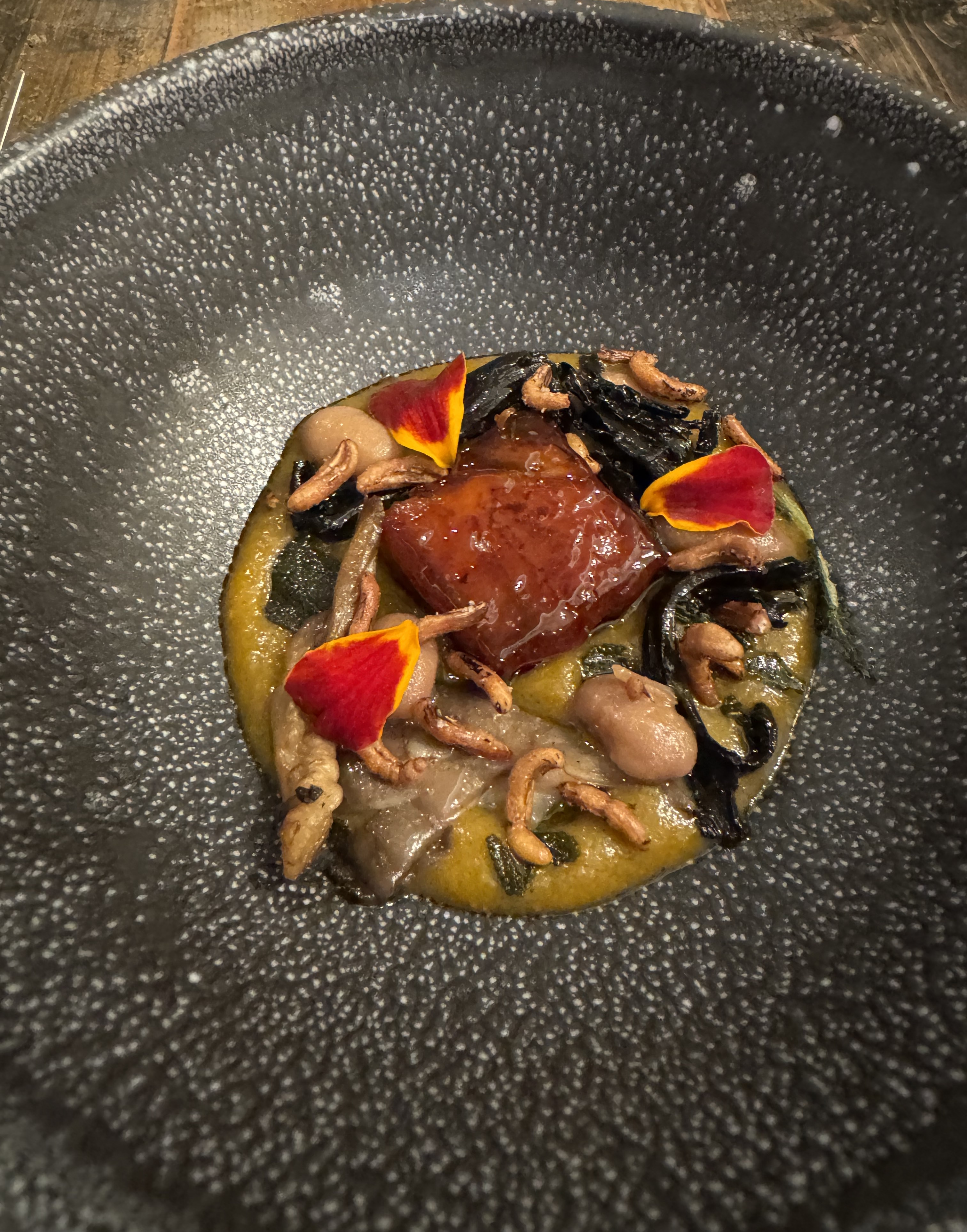 A very shallow black bowl. There is an orange sauce in it, dressed with a lot of toppings: shiny pale-brown cooked mushrooms, pinto beans, cooked seaweed and herbs. In the very center is a shiny red square of pork belly, in its candy shell.