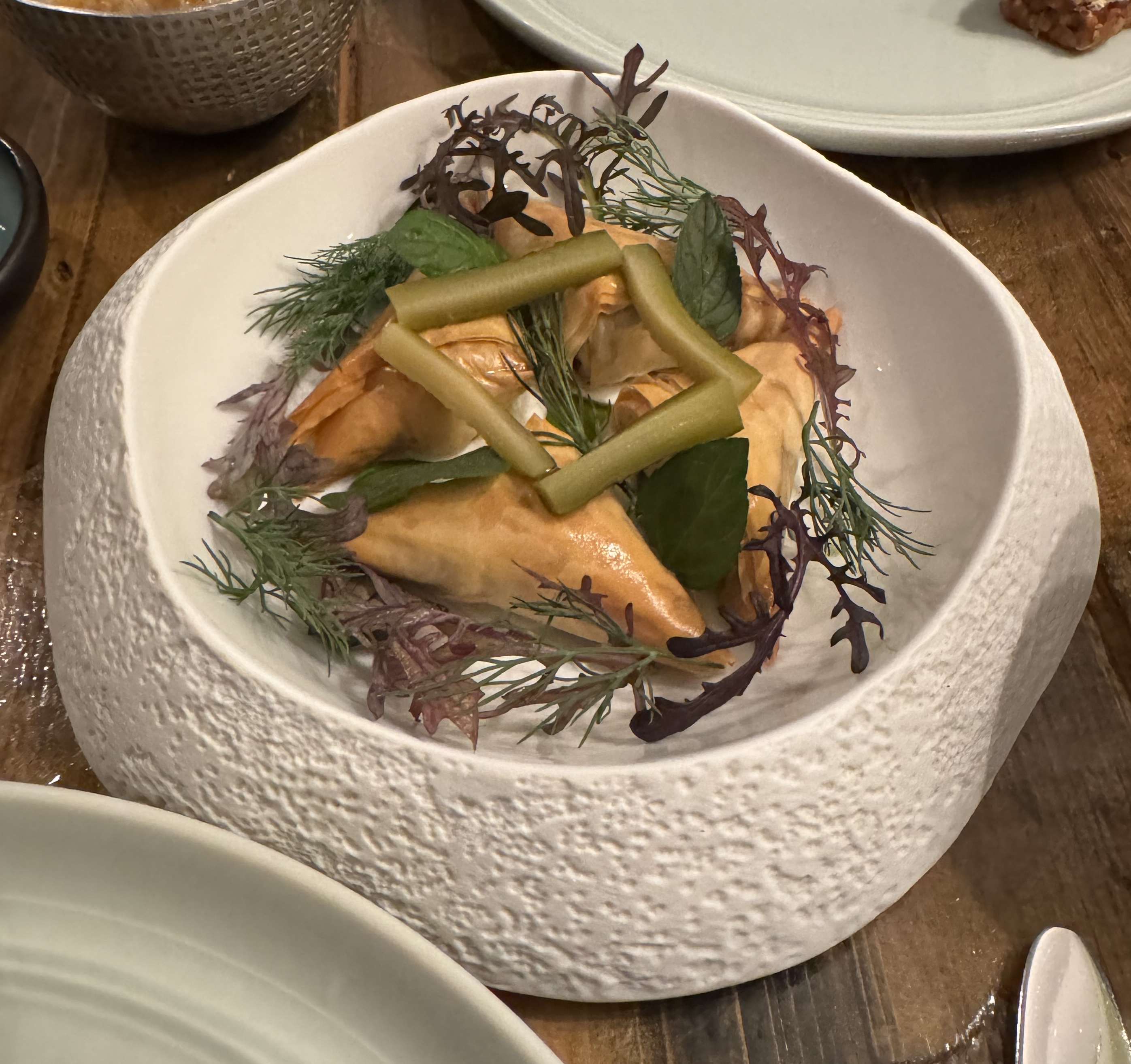 A wide, shallow bowl holding four triangular pastries. The pastries are placed so they form a square, and they are a beautiful golden colour. Around the pastries are dill sprigs and red microgreens. On top, as a bridge between each pastry, is a slice of dill pickle.