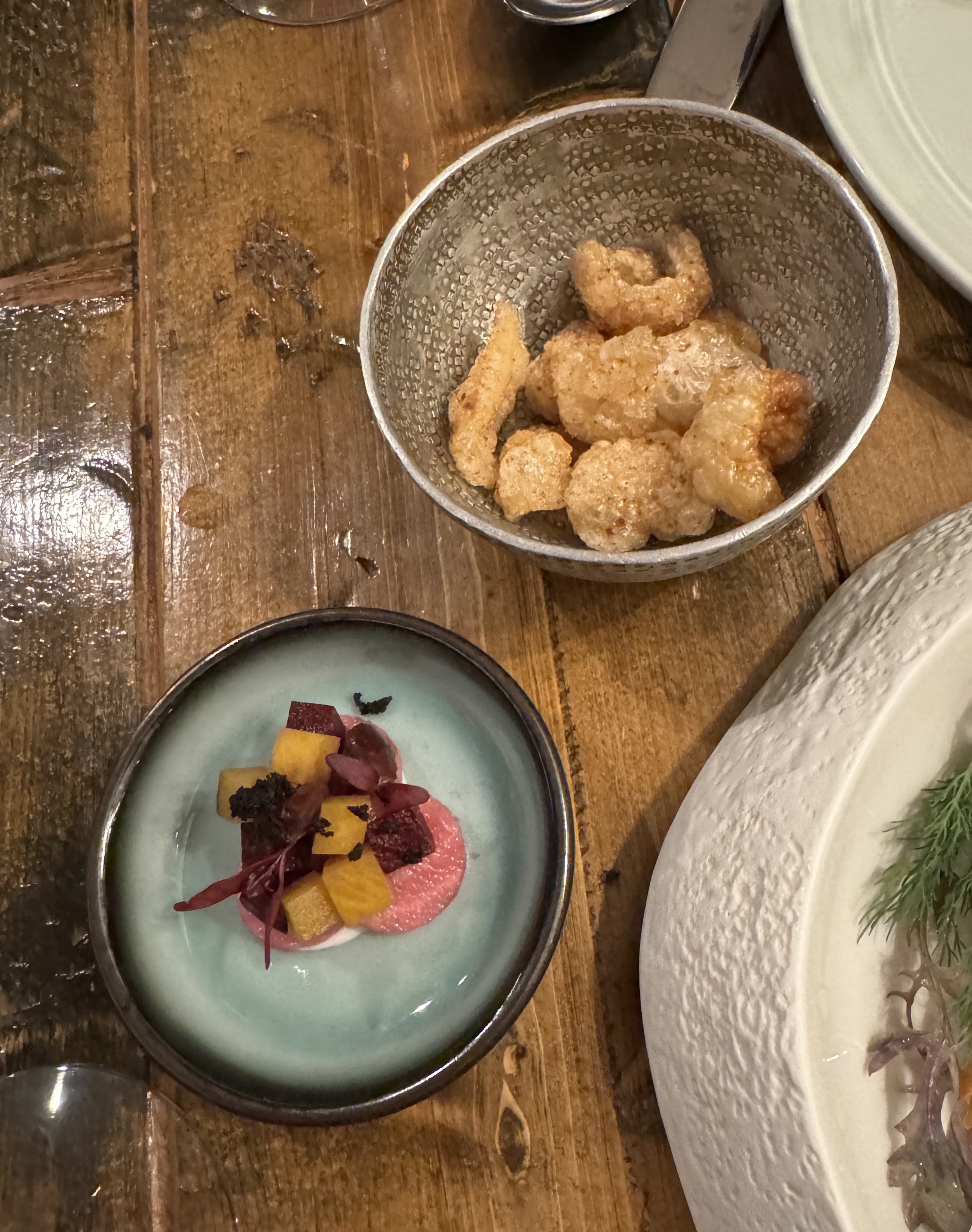 A small plate with a few red disks, topped with orange cubes and one red microgreen. Next to it is a bowl with fried chunks of pork skin, puffed up by the frying.