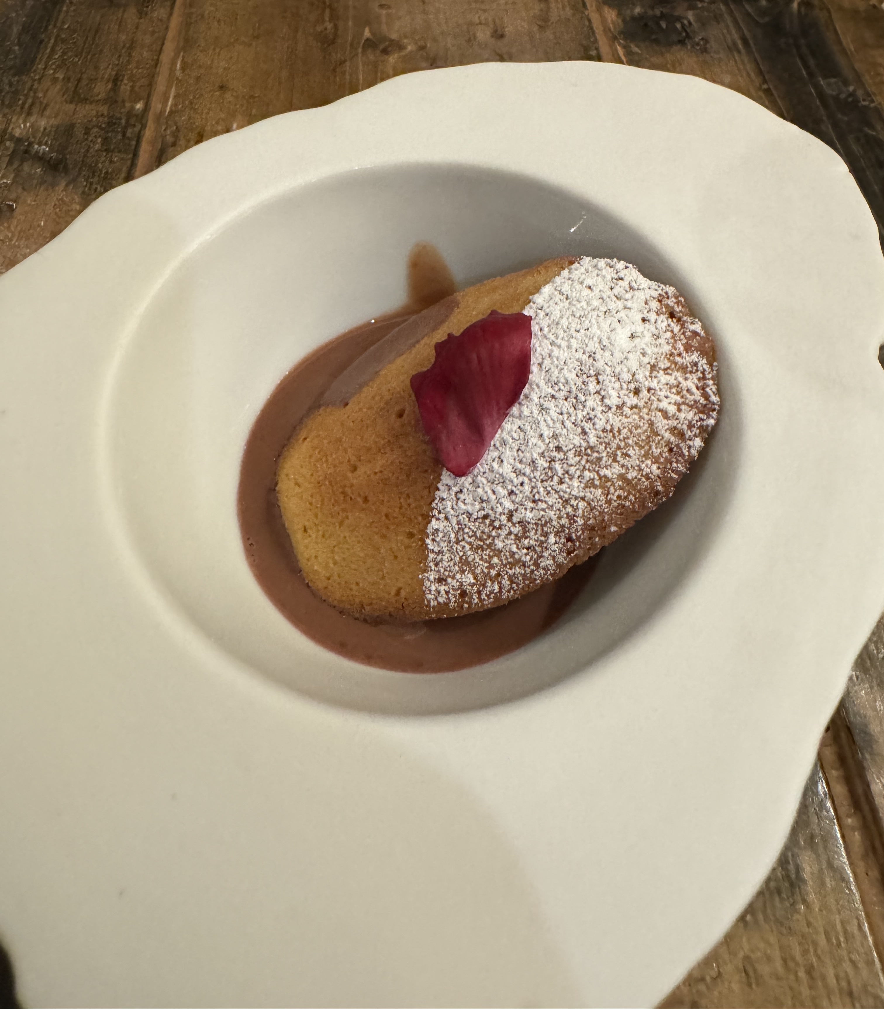 A bowl with a thin chocolate soup. On top of the soup is a Madeline-shaped cookie, golden brown, with one-half of it dusted in powdered sugar. The sugar has been applied precisely, giving it a clear line between the un-sugared cookie and the sugar. There is a red petal on the center as garnish.