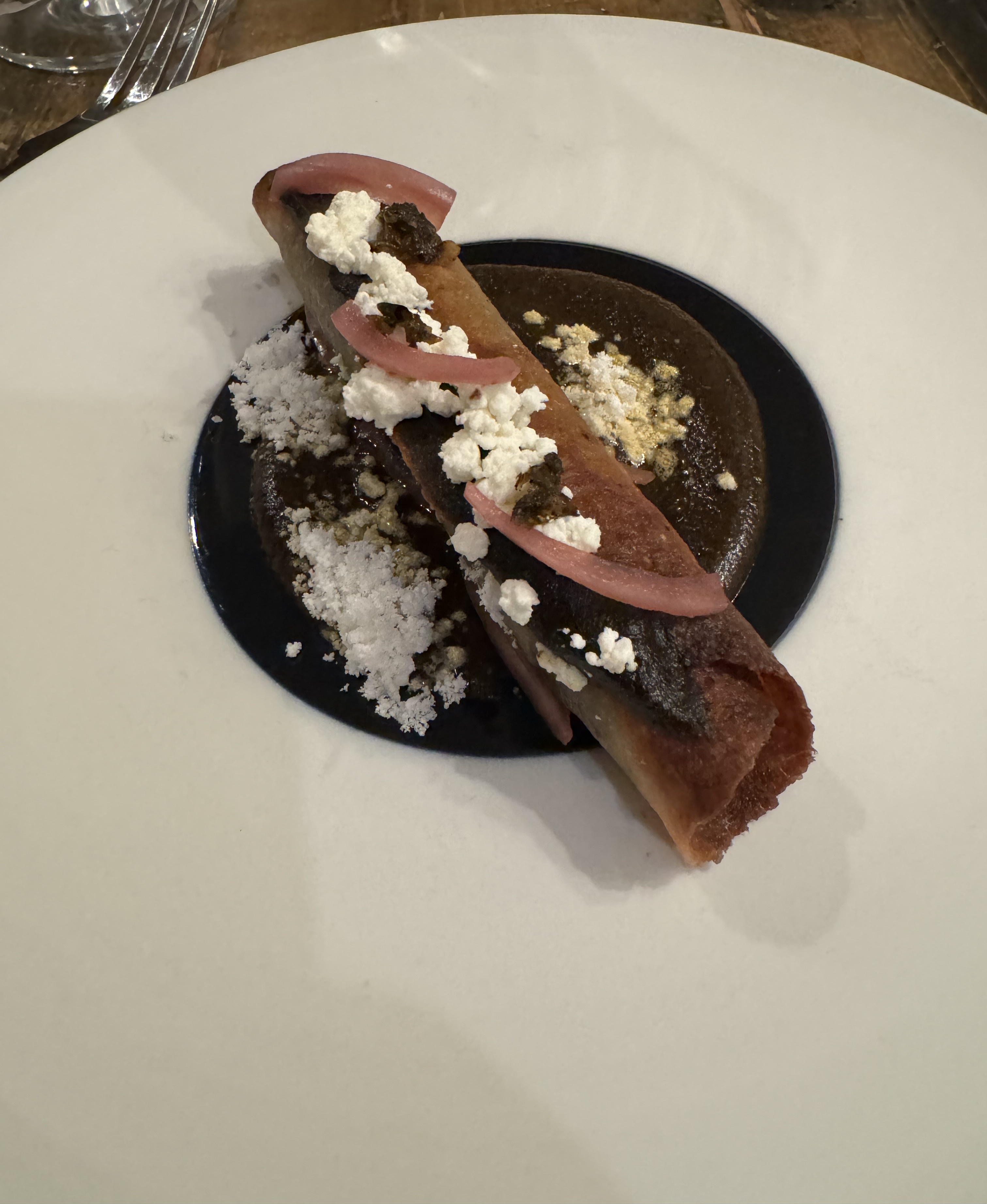 A circle of dark brown, chocolate-y looking sauce in the center of a plate. There is a rolled up tortilla, looking like a cigar, sitting in the middle of the sauce. Finely-grated white cheese is spread over it, and a few pieces of pickled red onion are carefully placed on top of the tortilla. The tortilla itself has been browned nicely after rolling it.