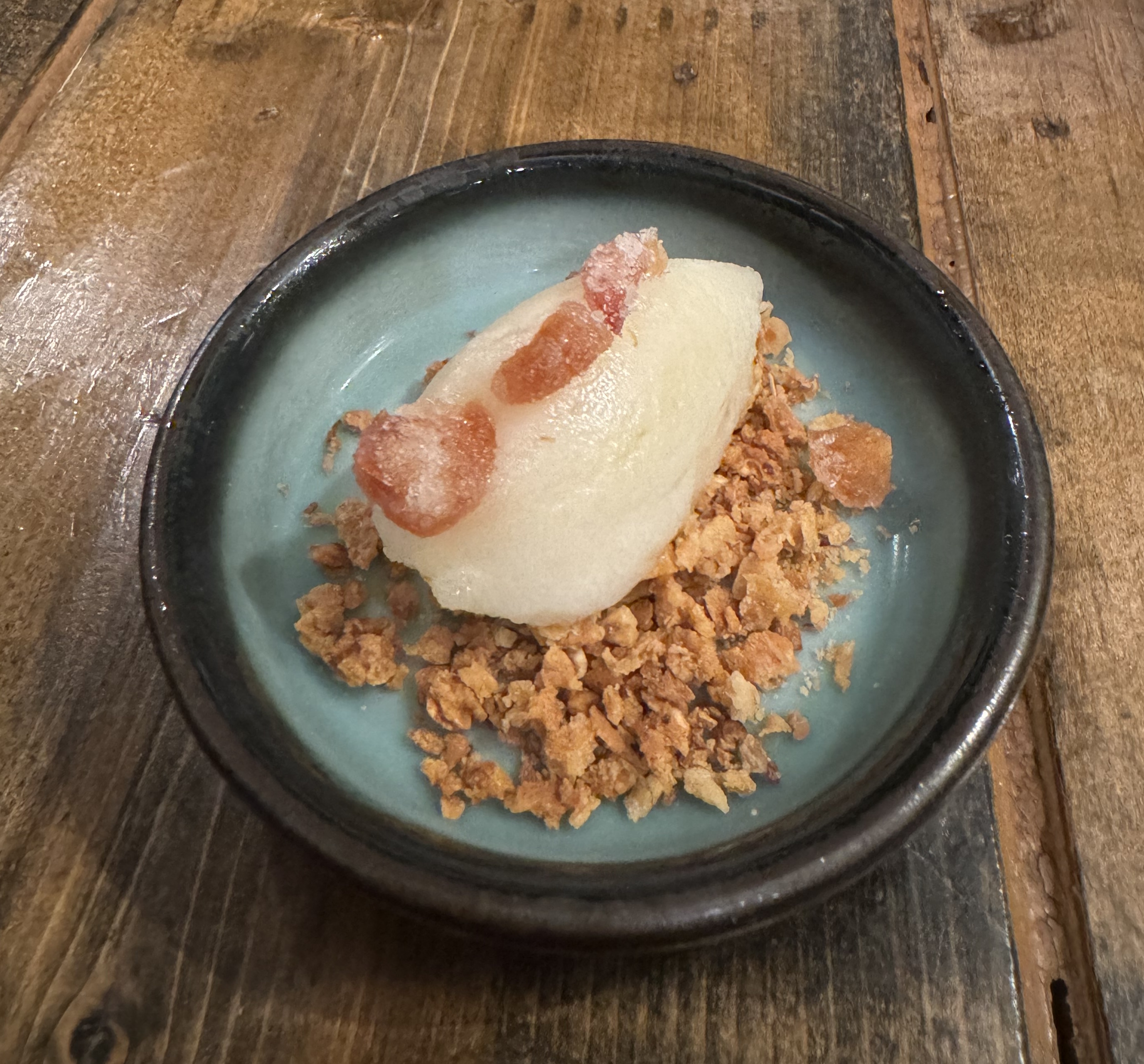 A quenelle of white sorbet sitting on top of toasted oats. There are three pieces of dark-orange candy on top of the sorbet, arranged in a line.