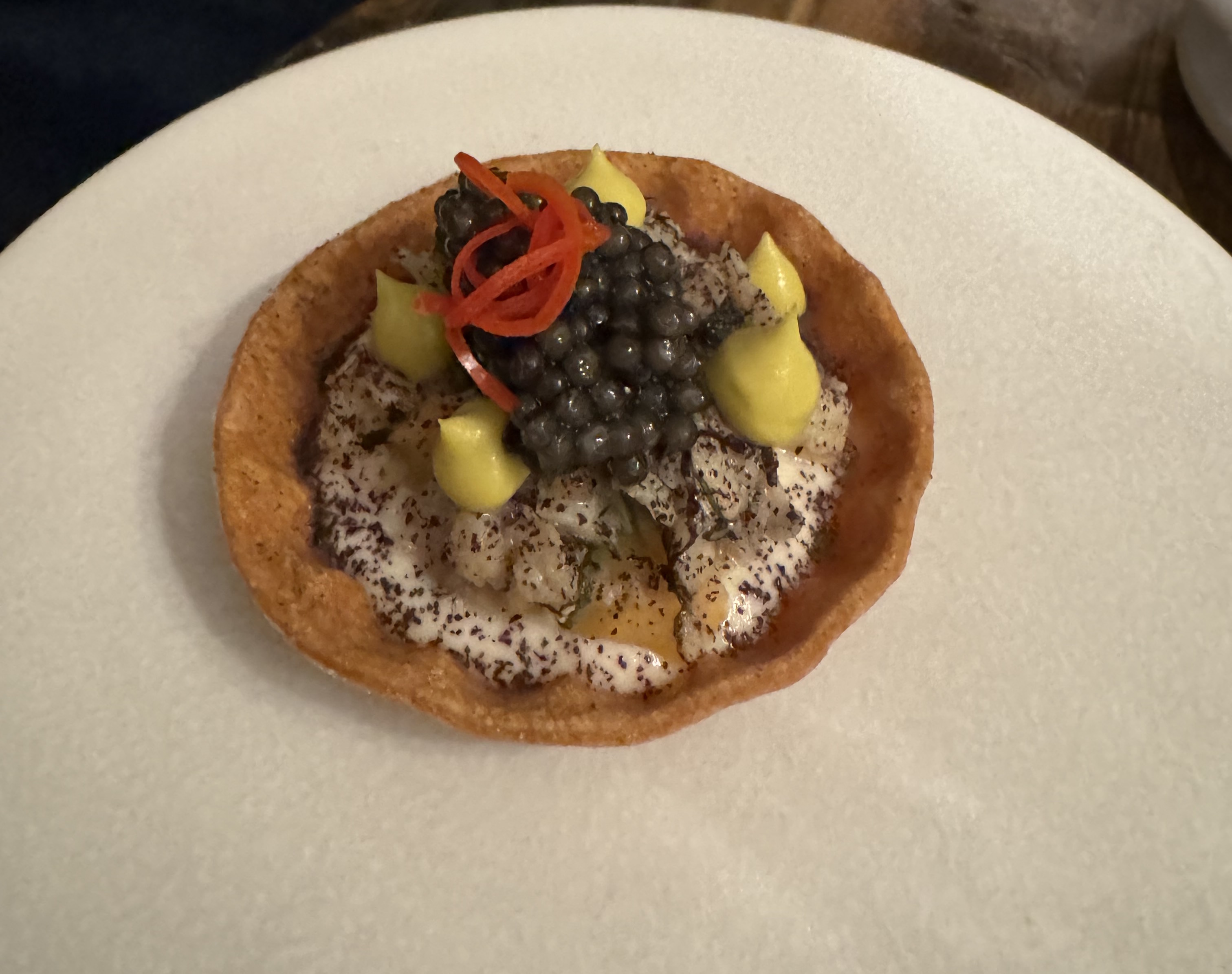 A wide, shallow tartlet shell on a plate. The shell is a dark golden colour. It is filled half-way with a white cream, which has been dusted with a red-ish powder. On top of the cream are five raindrop-shaped dollops of light yellow sauce. Between those is a mound of dark-green caviar, topped with a few strands of thinly-sliced red pepper.