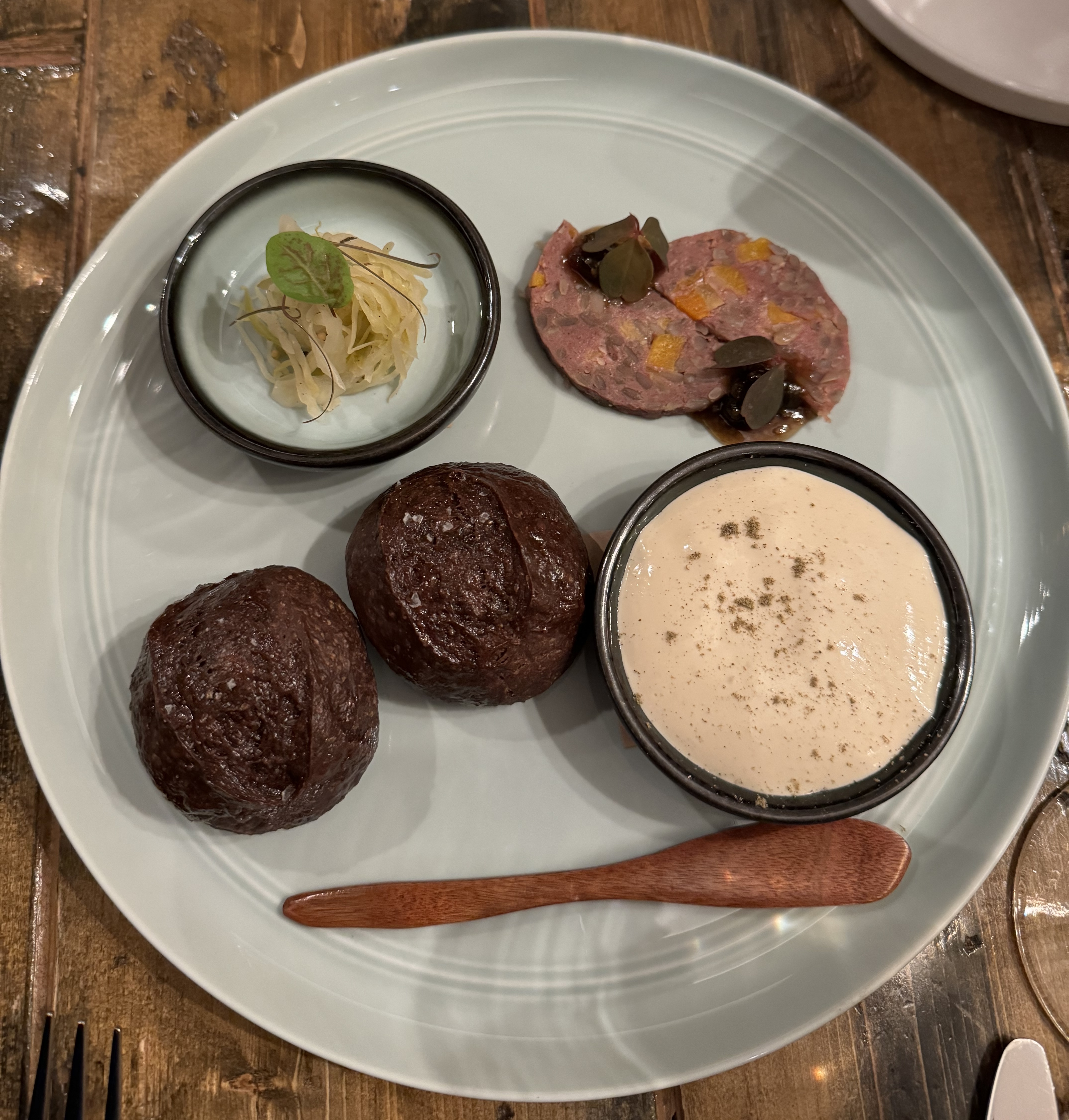 A plate with two dark brown bread rolls, and a round slice of meat cut into two half-moons and offset from each other slightly. There is a small dish of white, fluffy cheese, and another small dish with a mound of sauerkraut. A carved wooden paddle is on the plate, for spreading the cheese on the bread.