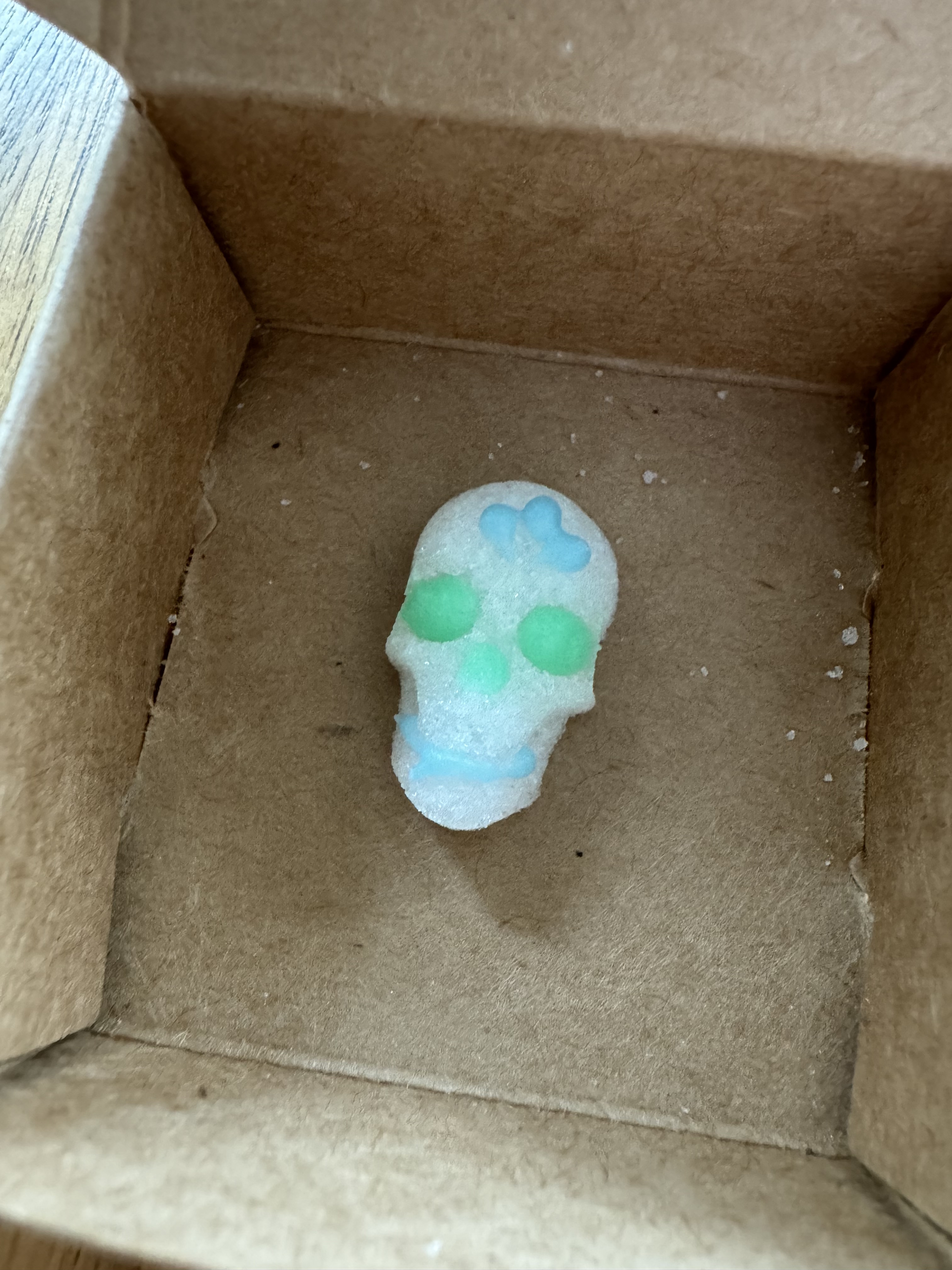 A tiny sugar skull in an equally-tiny box. The skull is white, with green eye sockets and nose socket. The mouth is blue, and there is a design with three pips on its forehead, also rendered in blue.