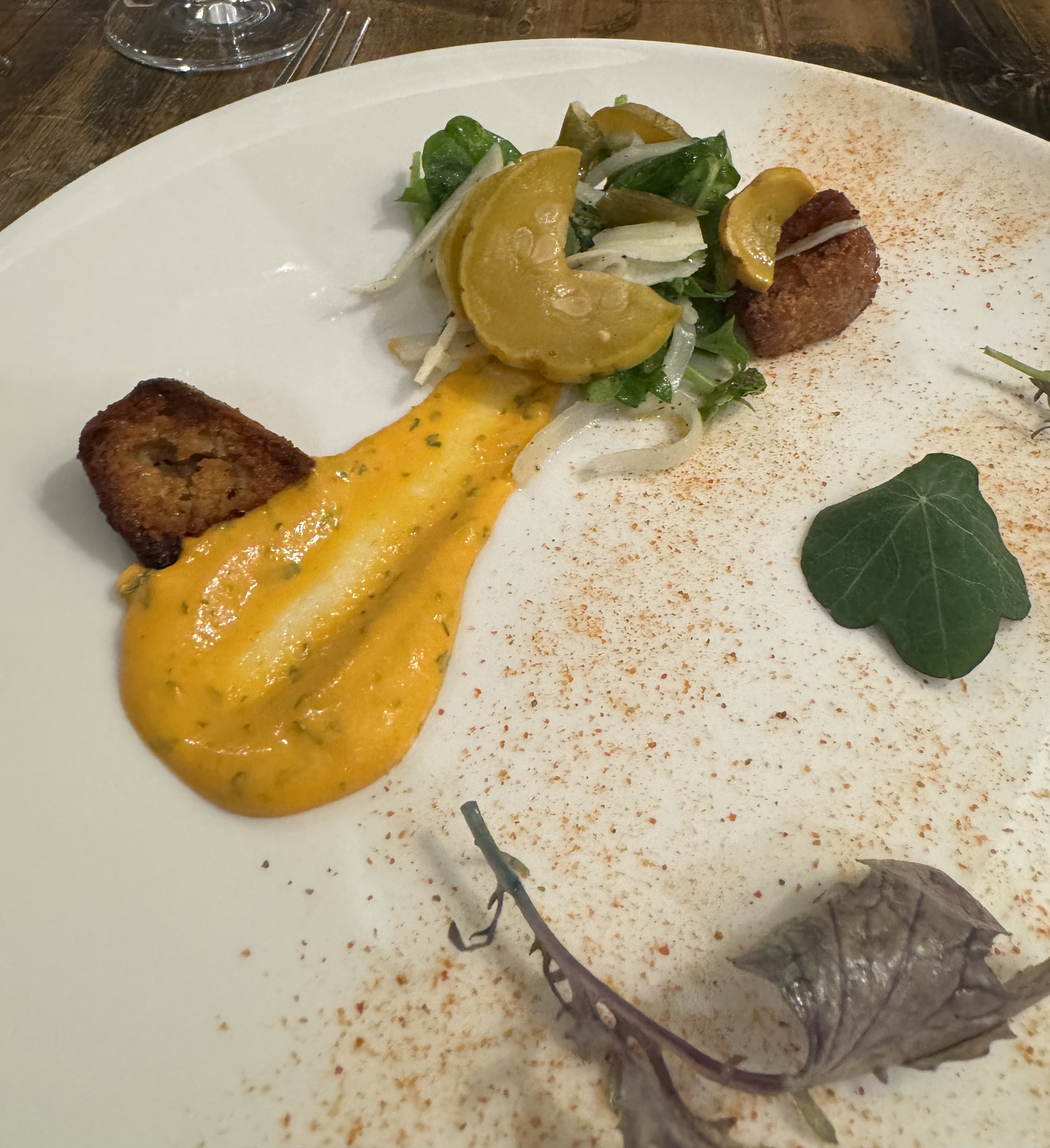 A deconstructed salad served on a large plate. There is a pile of lettuce and rounds of yellow squash. Extending out from that is a yellow sauce with hints of green herb flecks in it. The sauce is quite thick. Next to the sauce & salad are big crutons. There's a dusting of red powder to the side, and a few pieces of green & light purple lettuce scattered around the rest of the plate.