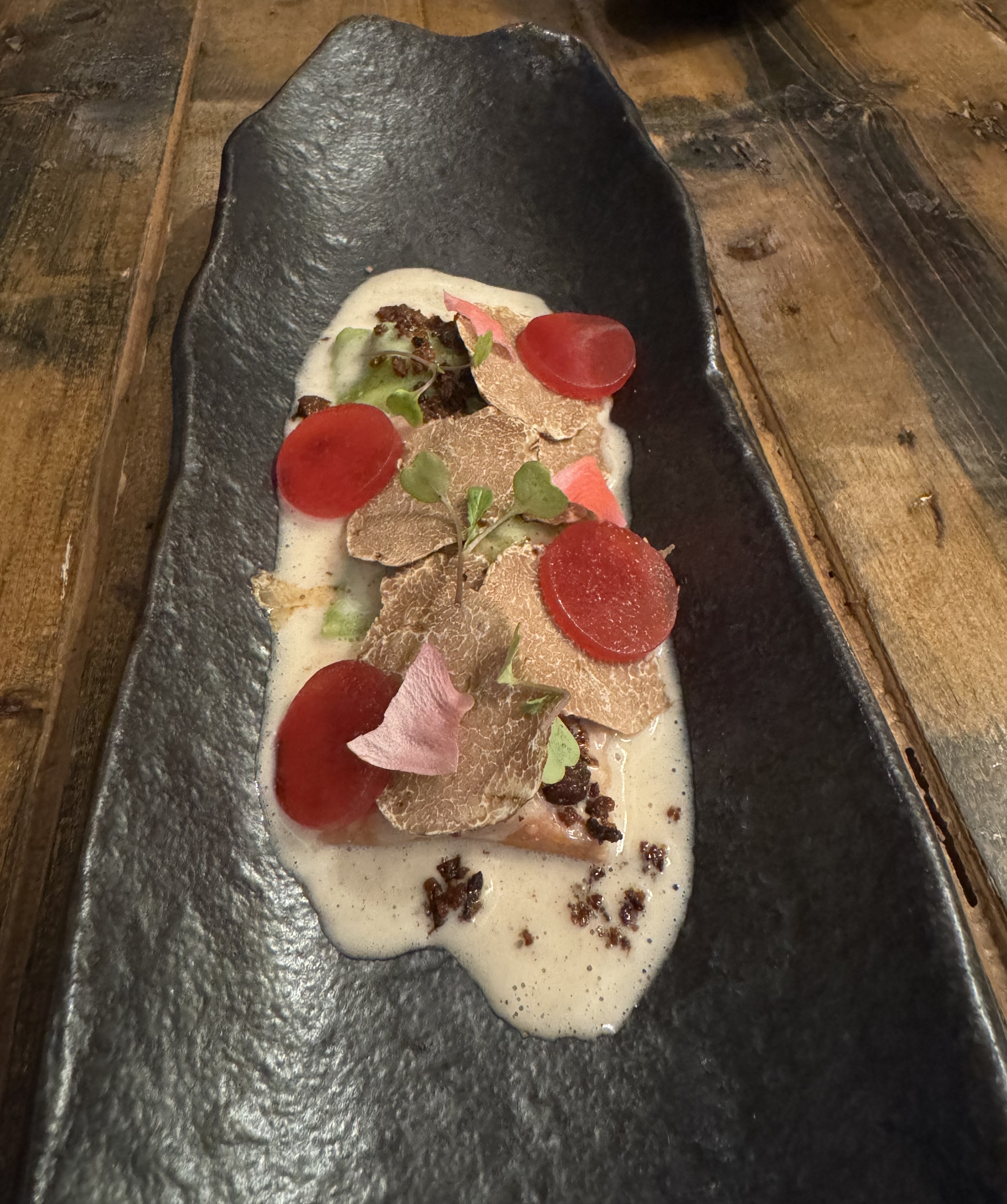 A shallow black trough dish, with pasta in a white sauce. The pasta is hidden underneath thinly-shaved white truffles, with only a few of the filled green pasta shapes visible. On top of everything are four discs of neon-red, a dark brown crumble, microgreens, and a single pink petal.