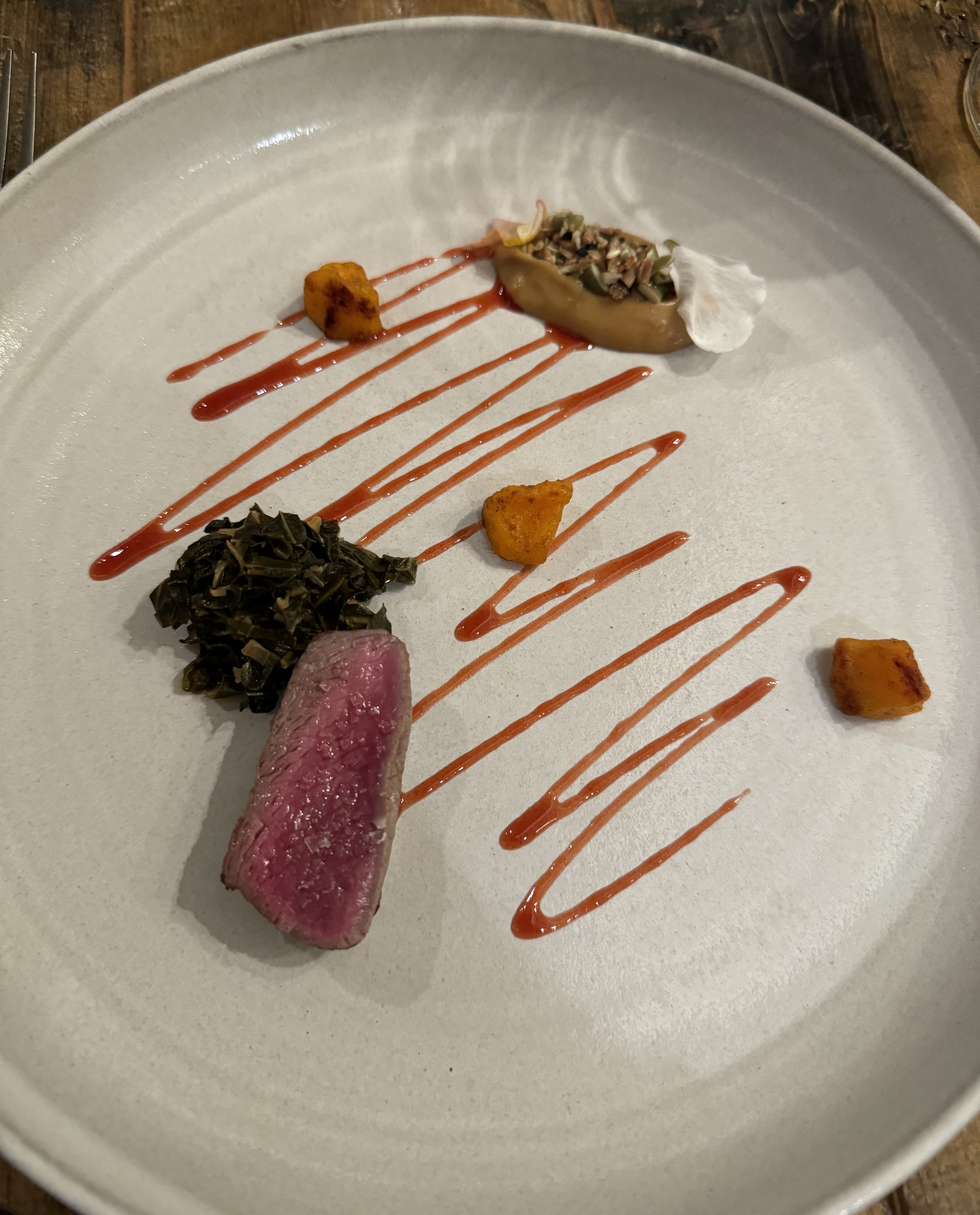 A large white plate. There is a single cut of venison, cooked rare, plated so the cut side is facing up and showcasing the pink meat. There is a pile of cooked, dark greens next to it. The center of the plate has squiggles of red sauce, and on the far side of the squiggle is a quenelle of yellow sauce with puffed rice on top. There are a few chunks of orange potato, with some charring, placed randomly between the meat & quenelle.