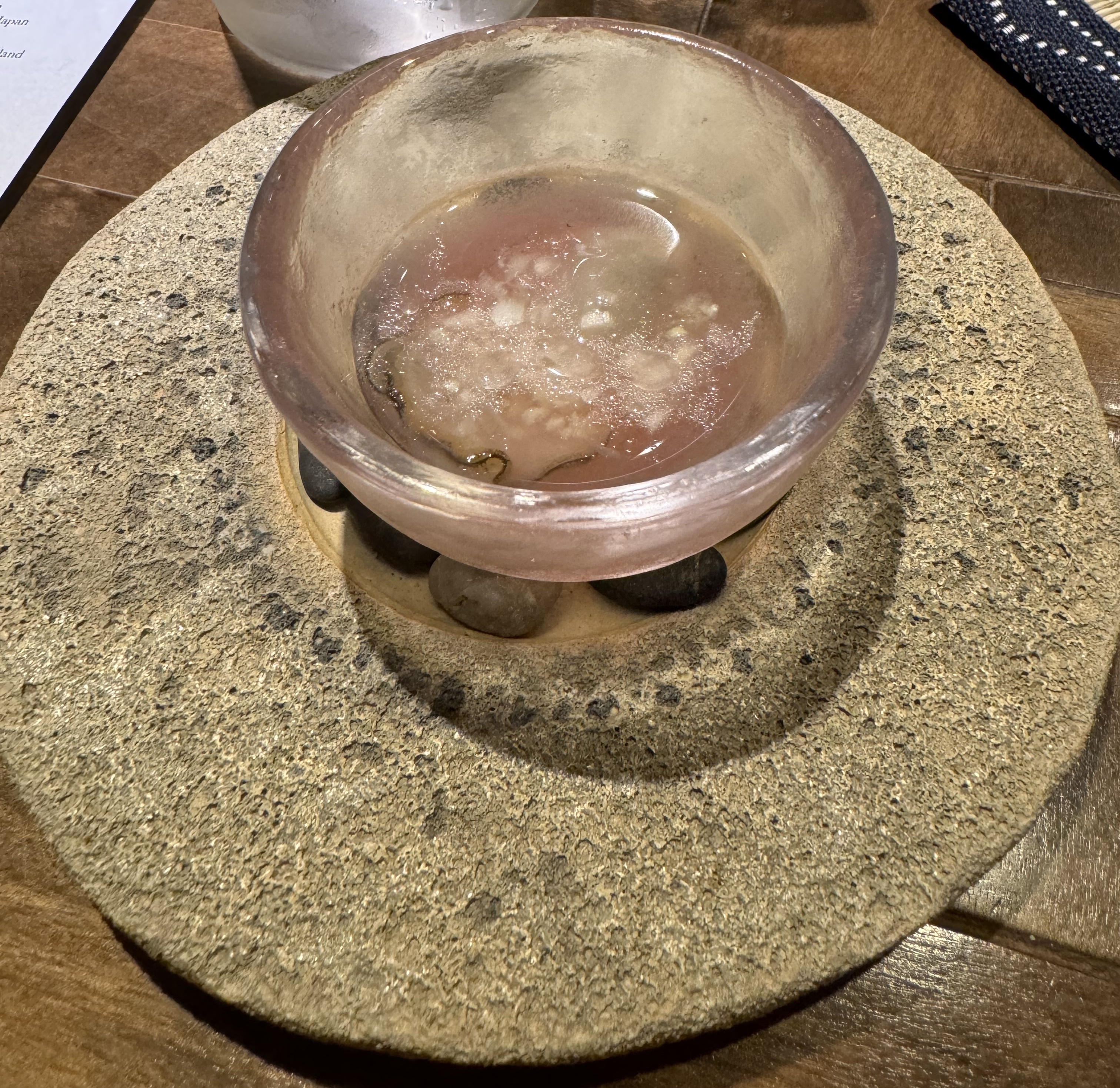 A thick, translucent pink bowl sitting on top of a few smooth stones. There is fluid in the bowl, and in the bottom, you can make out a white oyster meat fringed with black.