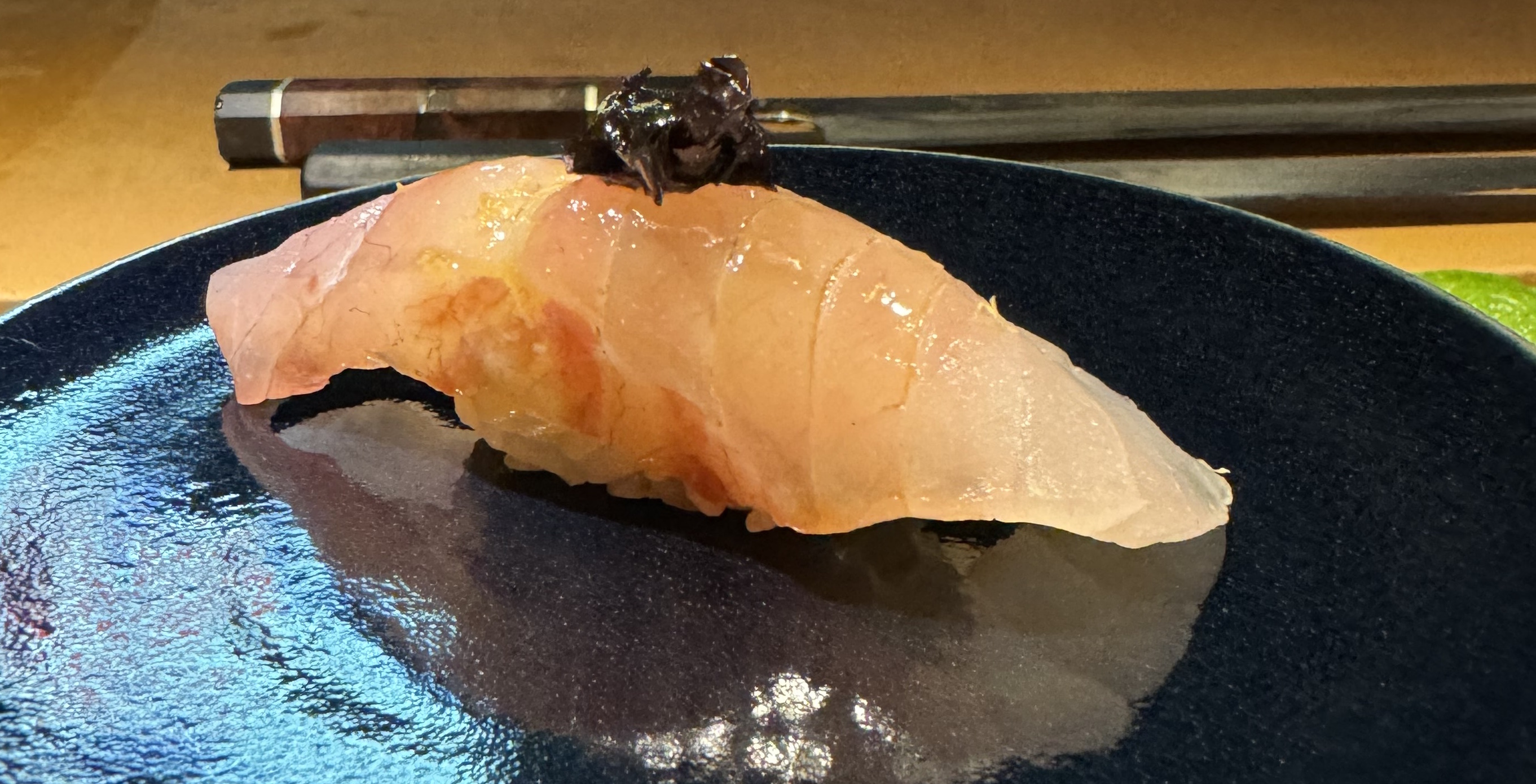 A piece of slightly-pink white fish sitting on top of rice. The fish has been very lightly brushed with a dark sauce down the middle; the ends are the original colour, but the center of the fish glistens with the sauce. There is a small heap of black shredded seaweed on top as garnish.