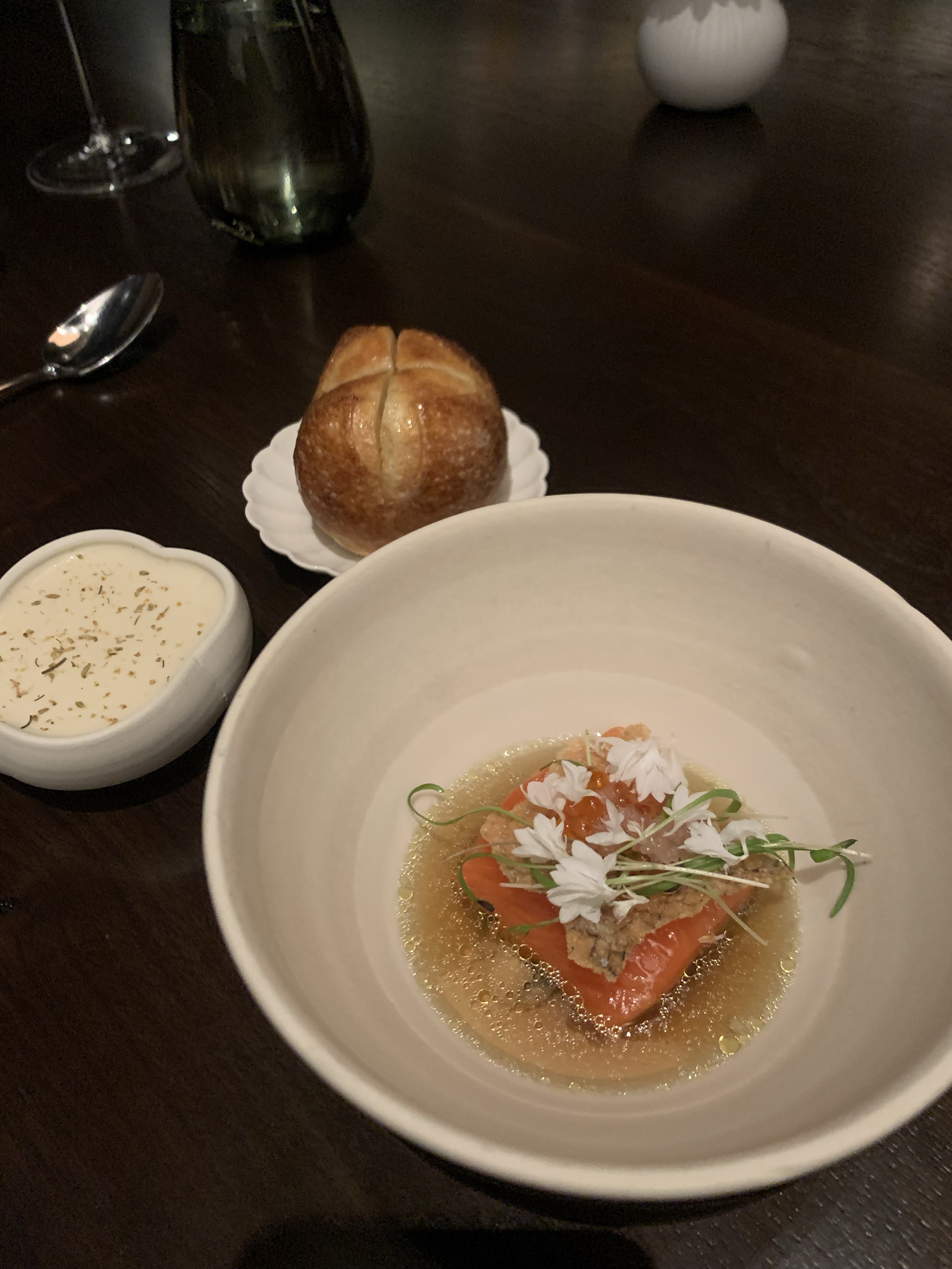 Dinner roll next to a bowl of with an orange trout fillet, broth, and skin puffed & placed on top of the filet
