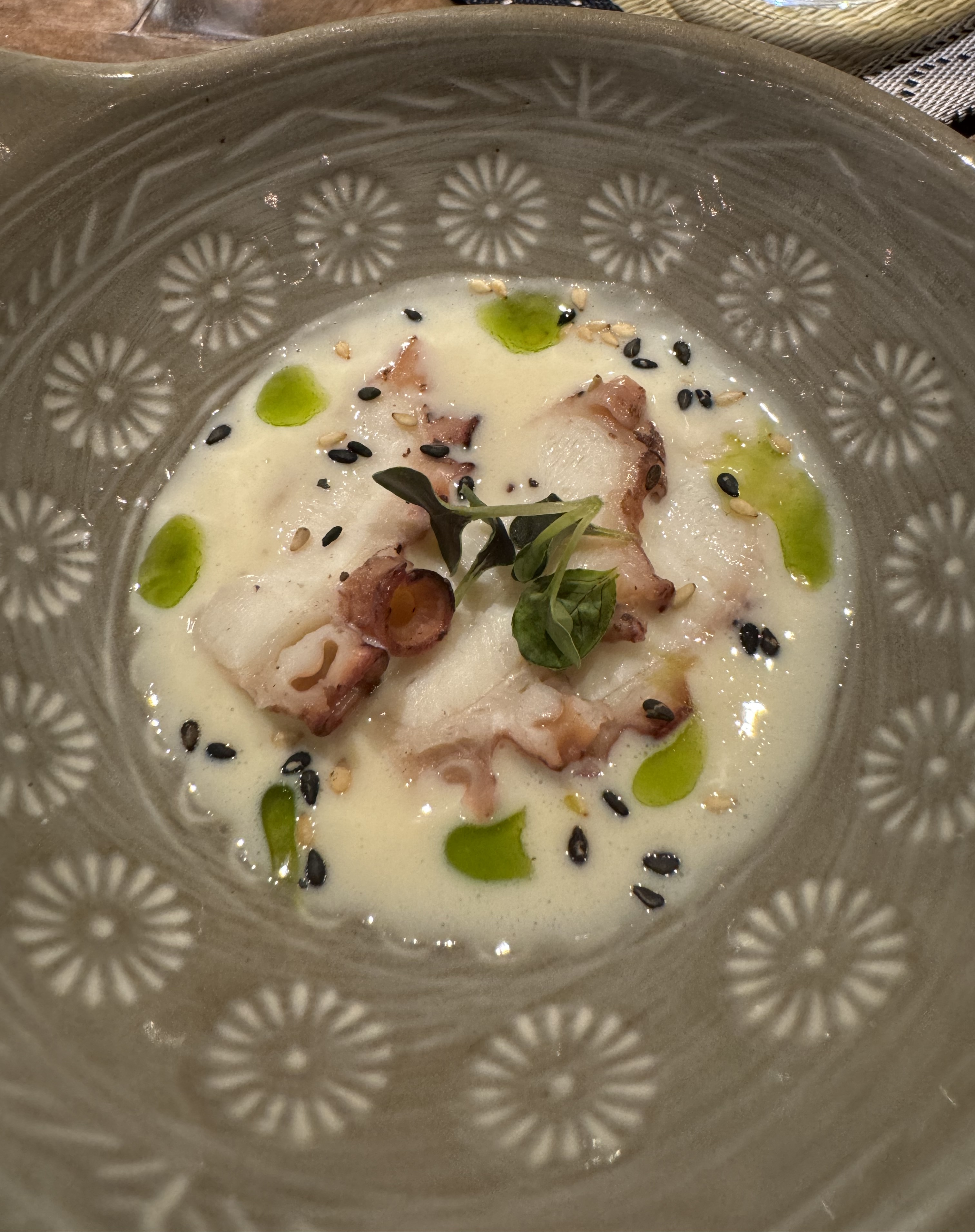 A bowl with a white broth speckled with dots of bright green oil. There are slices of white octopus meat in the broth, partially submerged. The meat is fringed with the dark pink hue of octopus skin.