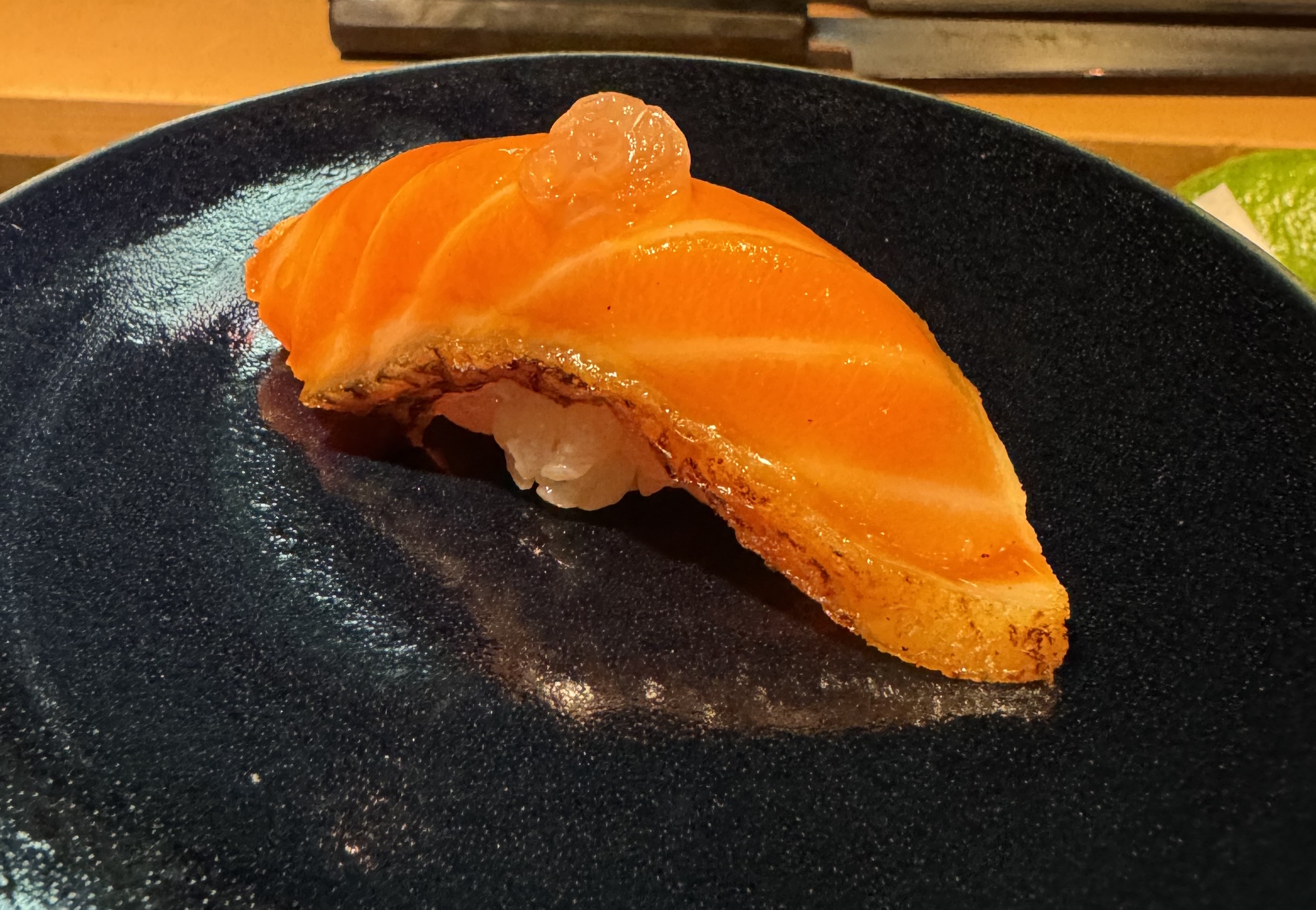 A piece of orange fish with white stripes of fat sitting on top of rice. There is a thin band of skin, which is similar in colour to the fish, but has some charring. There is an orange pickled sphere as garnish on top.