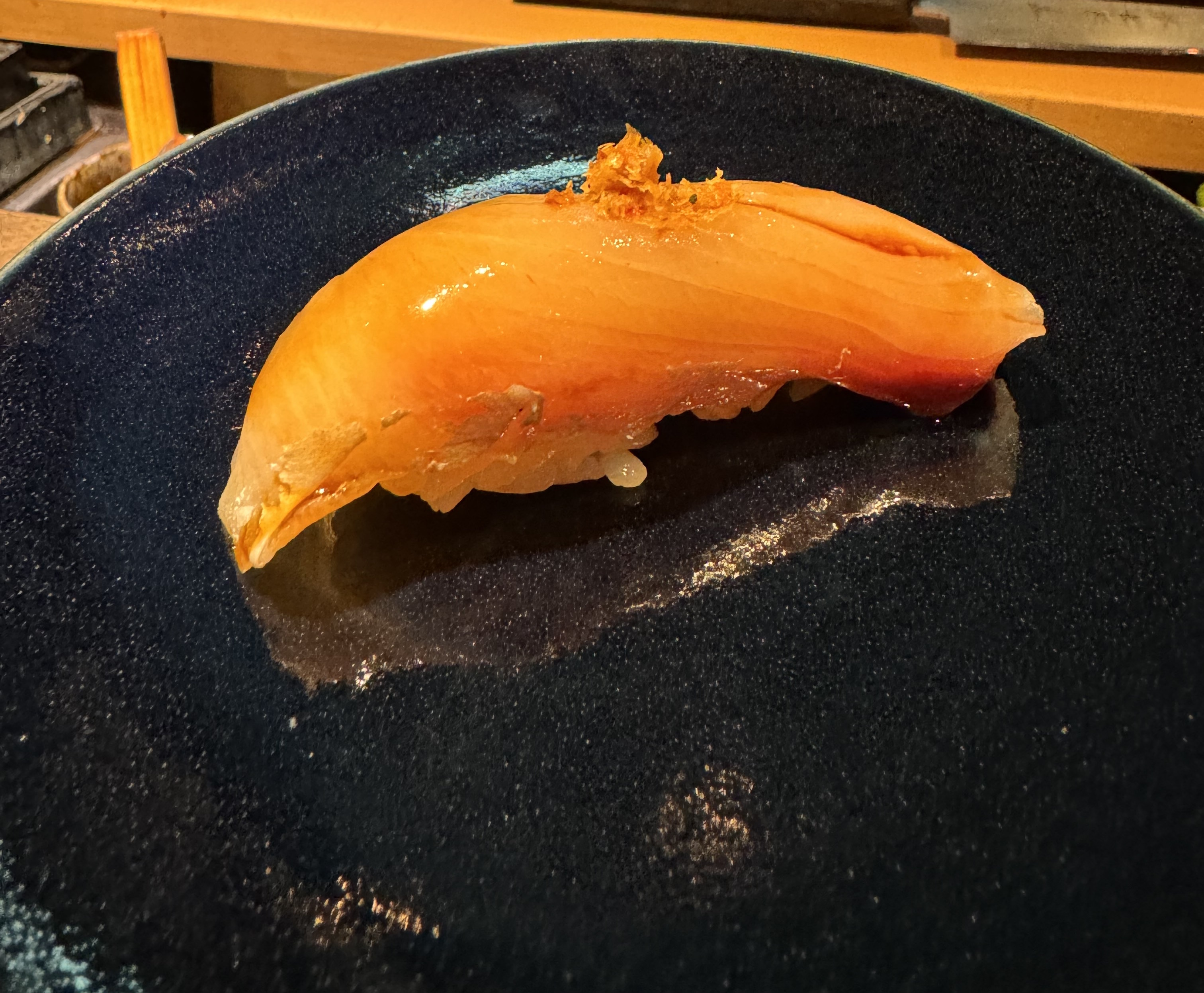 Piece of orange fish on top of rice. There is no prominent fat banding, but you can see the separation between the layers of the flesh. It's has a sheen of being brushed with oil. There is a powder in a pile on top that is red, yellow, and a speck of green.