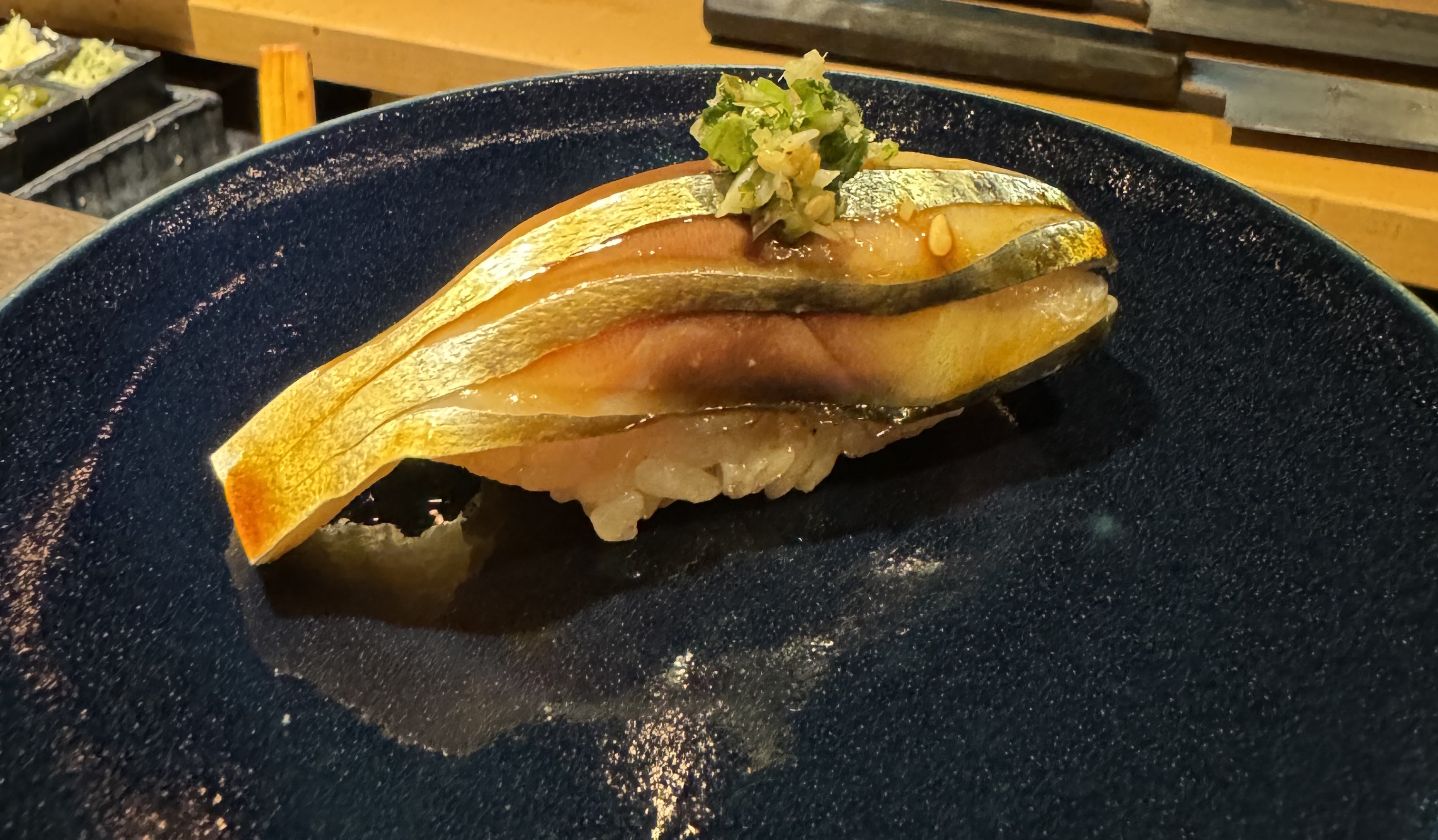 A single piece of fish sitting on top of rice. The fish is skin-side up, and it has silvery skin. The piece has been cut and fanned out, so you can see a band of skin and the flesh beneath, stacked up. The flesh itself is pale pink, with a dark spot near the skin on one of the layers. It has been brushed with a dark sauce before cutting, giving the skin a burnished look. There is a pile of green onion on top, and a bit of sesame seed.