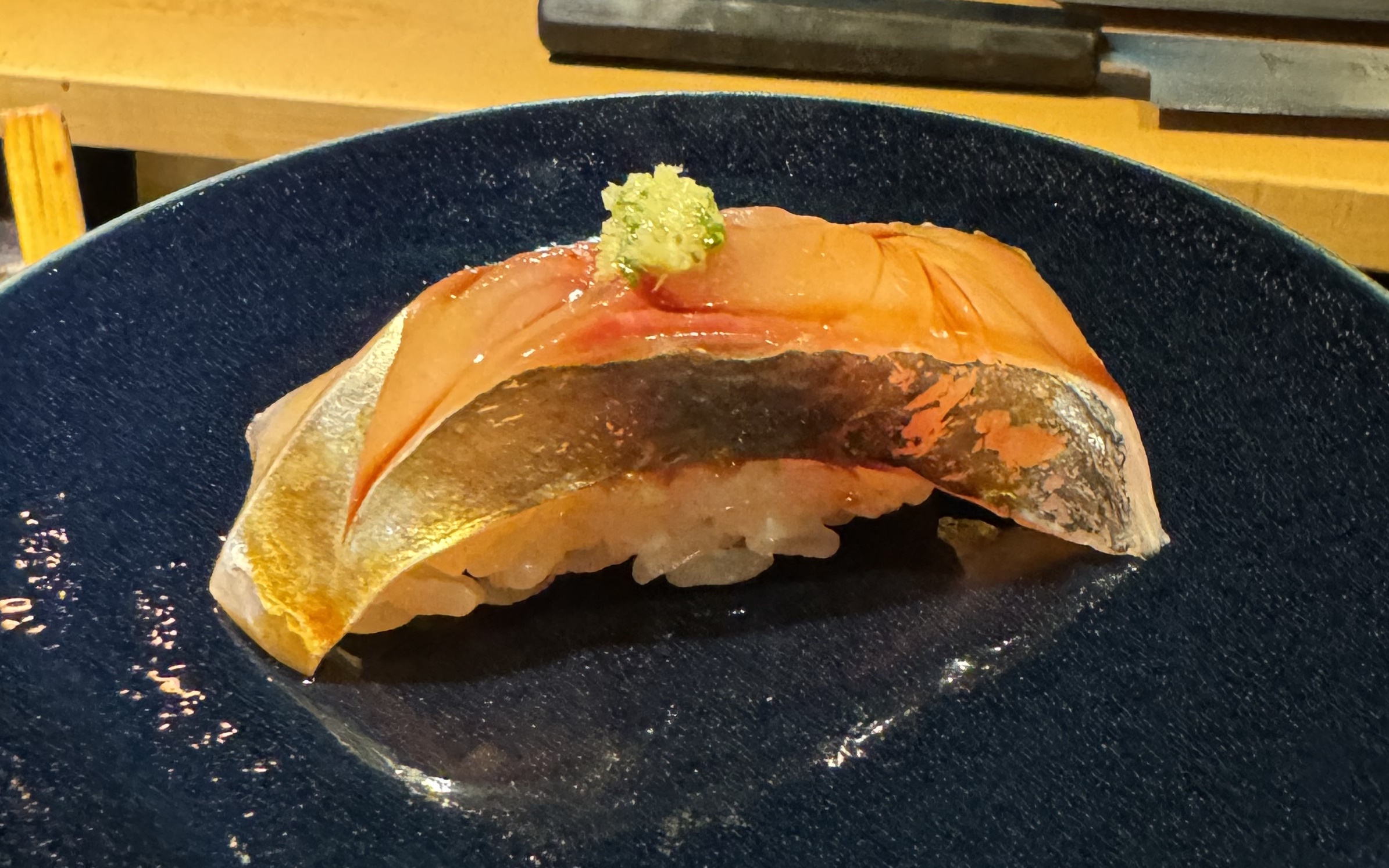 A piece of fish sitting on top of rice. The fish is pale, brushed with a dark sauce, giving it a deep golden hue. Sauce is pooled in some of the valleys in the flesh. There is a thick band of silver skin along the side. There is a small pile of grated green wasabi on top.