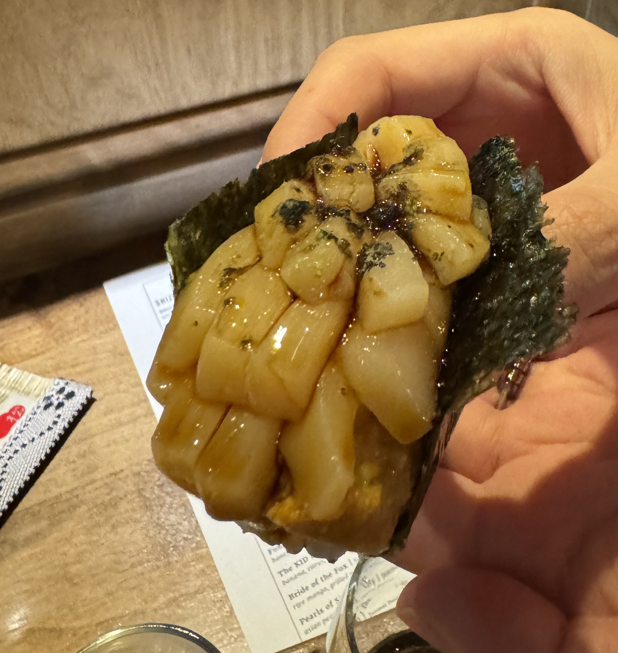 White scallop meat that has been sliced into rectangles and stacked up inside a piece of nori, as a hand roll. The scallops have been brushed with a dark sauce, giving them a mahogany hue. Someone is holding the handroll up to the camera, since it would not stay together if put on a plate.