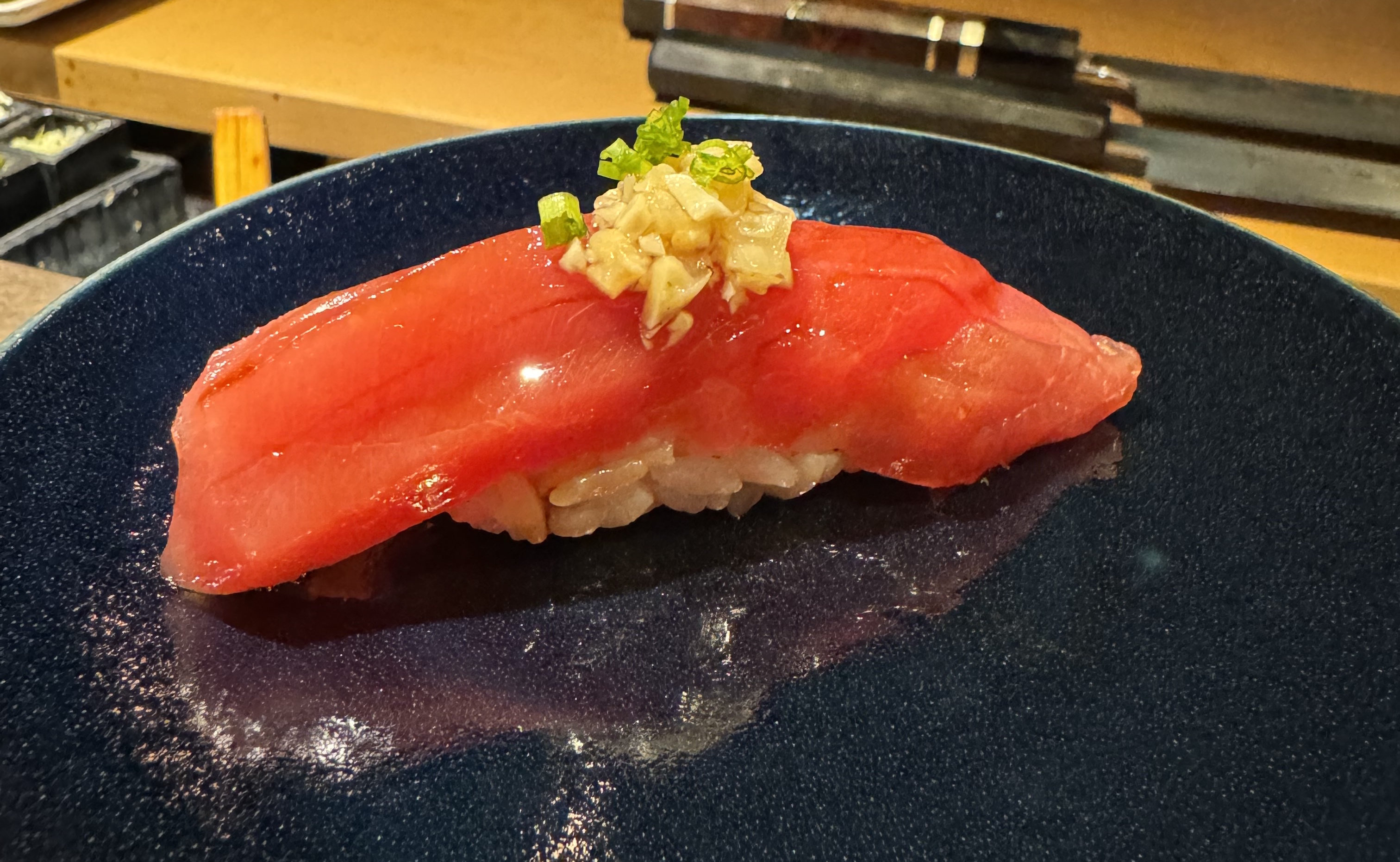 A red piece of fish on top of rice. The fish is bright red, with some bands of slightly-lighter-hued flesh. It glistens in the light. There is chopped yellow ginger on top, and some tiny rounds of green onion.