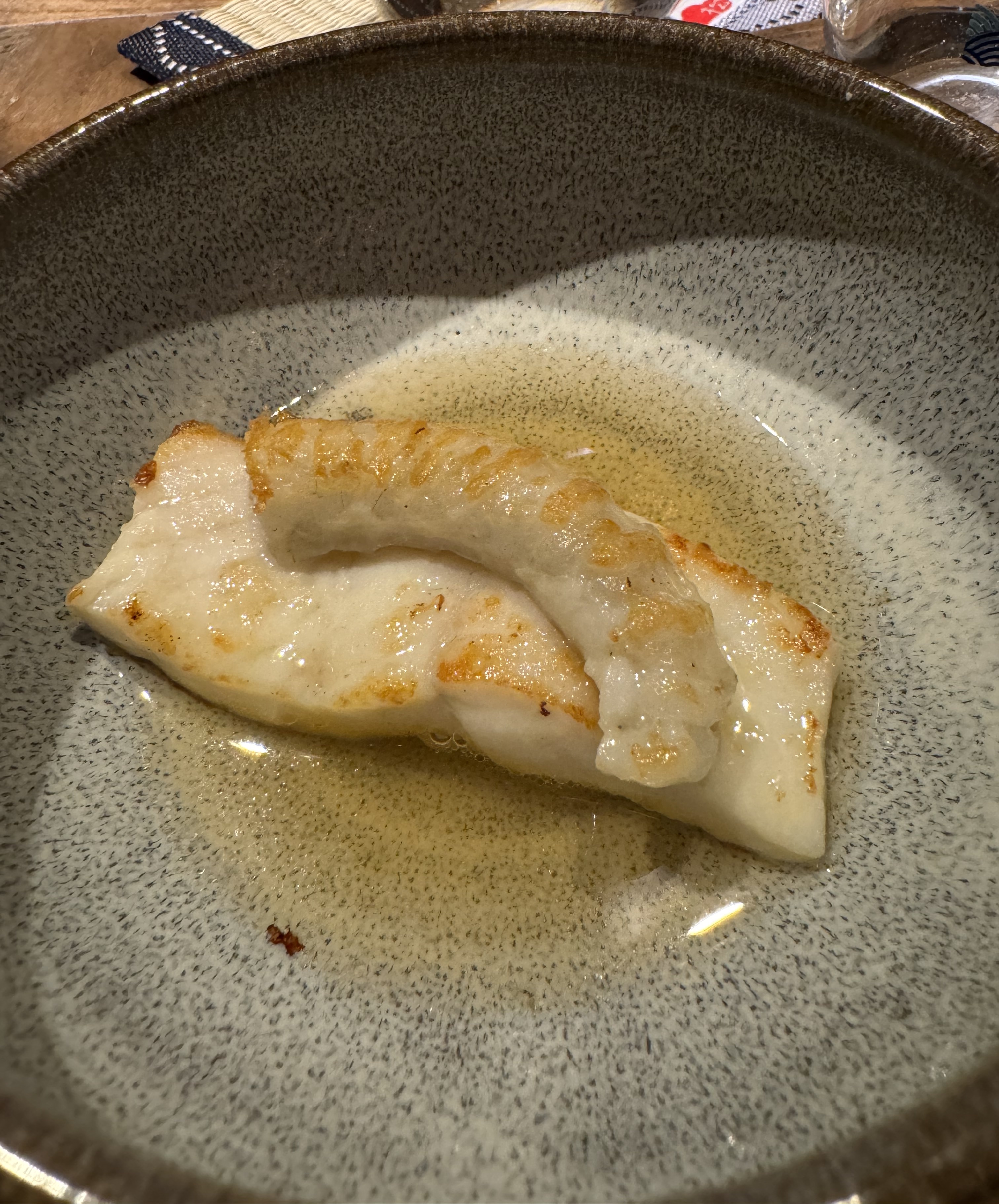 A bowl with a shallow pool of golden broth at the bottom. There are two pieces of cooked white fish: one a rectangular filet, with the center being thicker than the ends, and the other a curved piece of slightly-greyer meat. Both pieces have some golden brown charring around the edges. 