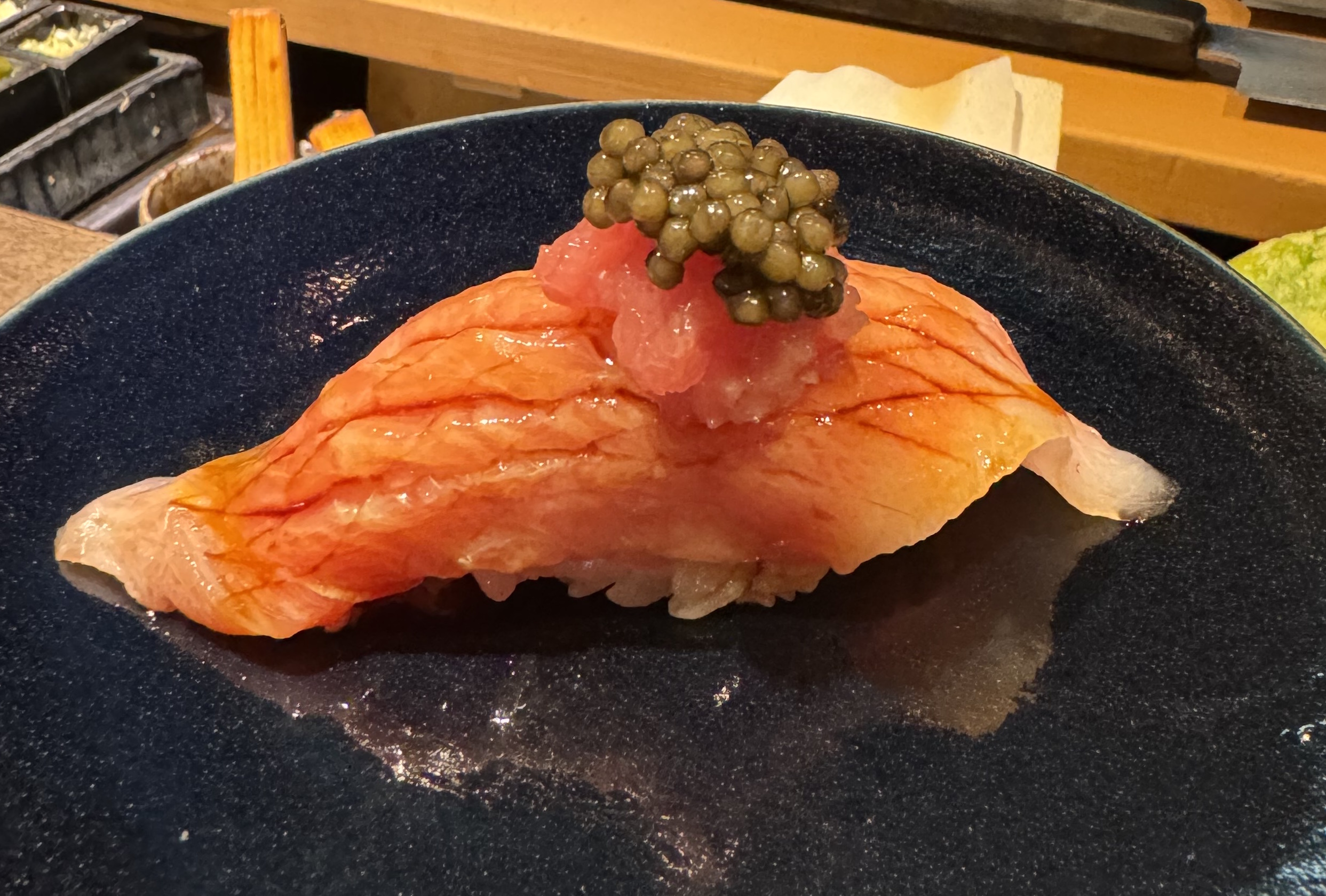 A light pink slice of fish on top of rice. The very tips of the fish are pale pink; the rest has been brushed with a sauce, which brings out the redness of the flesh. The sauce pools in slices made into the fish. There is a heap of red minced tuna on top, holding up a mound of green caviar.