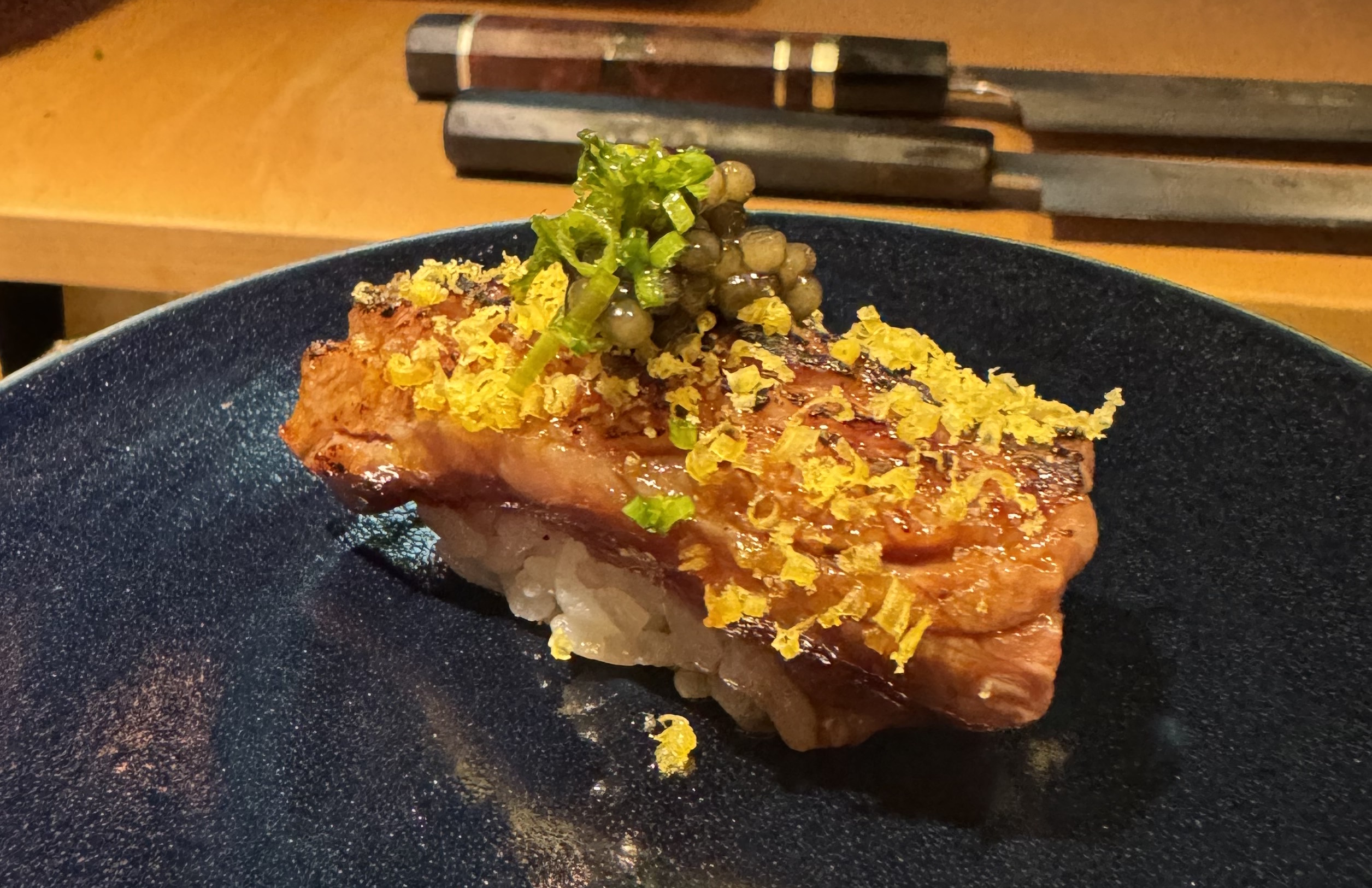 A lumpy piece of beef atop rice. It's rectangular, and has some charring in spots, but is otherwise pink. There is grated yellow stuff over the beef, and in the center, there is a heap of dark-green caviar spheres topped with some tiny rounds of bright green onion.