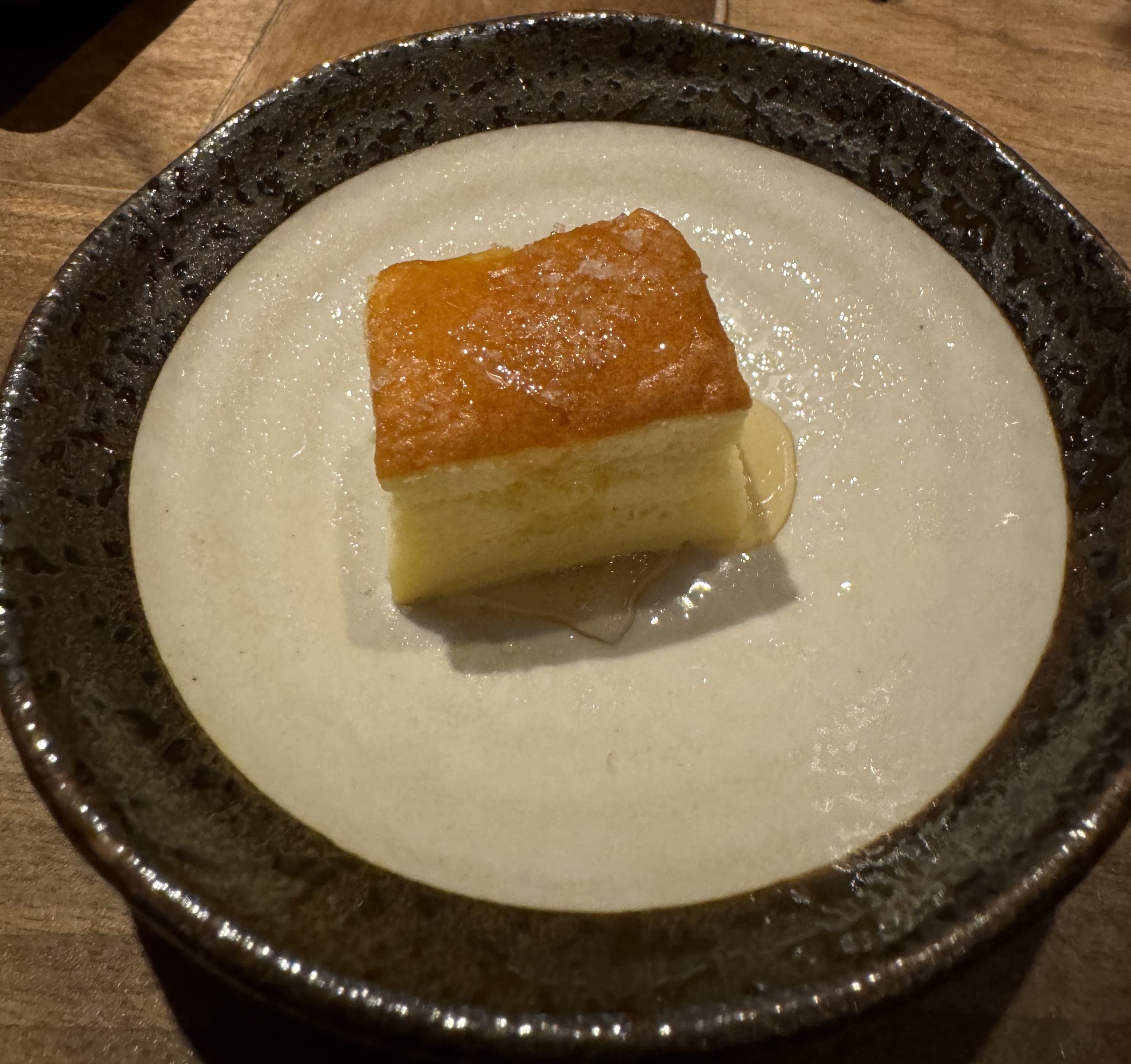 Plate with a square piece of cake. The cake is light yellow and very fluffy. The top is dark brown. There is a little bit of honey-coloured liquid around the bottom.