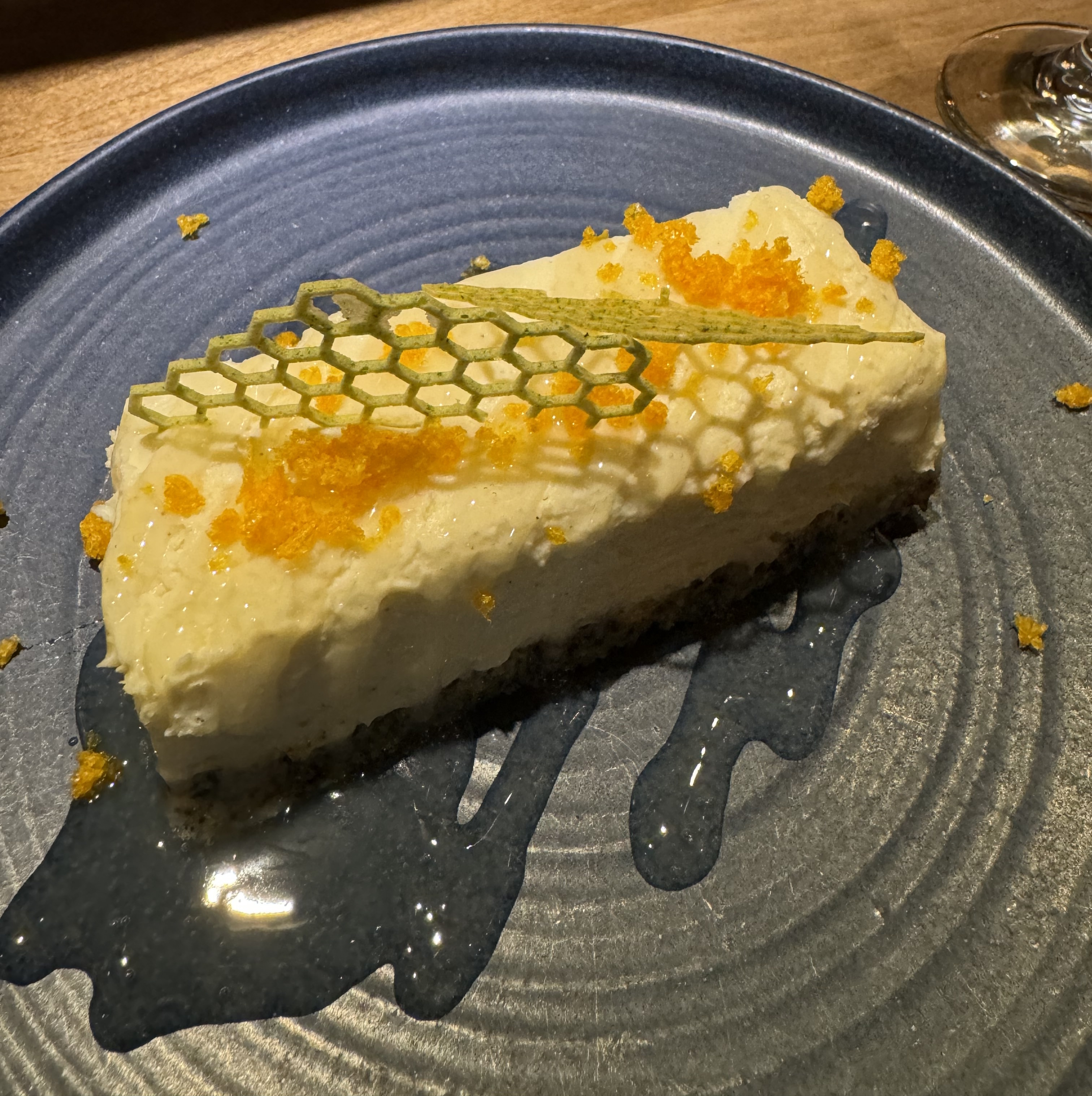 A bar of cake on a plate, with honey-coloured sauce around the base. The cake has a dark cookie-crumb base, with a stable white custard atop it. The custard generally holds its shape, but looks gooey instead of giving you a clean break when you cut it. The top is garnished with a cookie shaped like a honey-comb, and some grated dark yellow lemon peel.