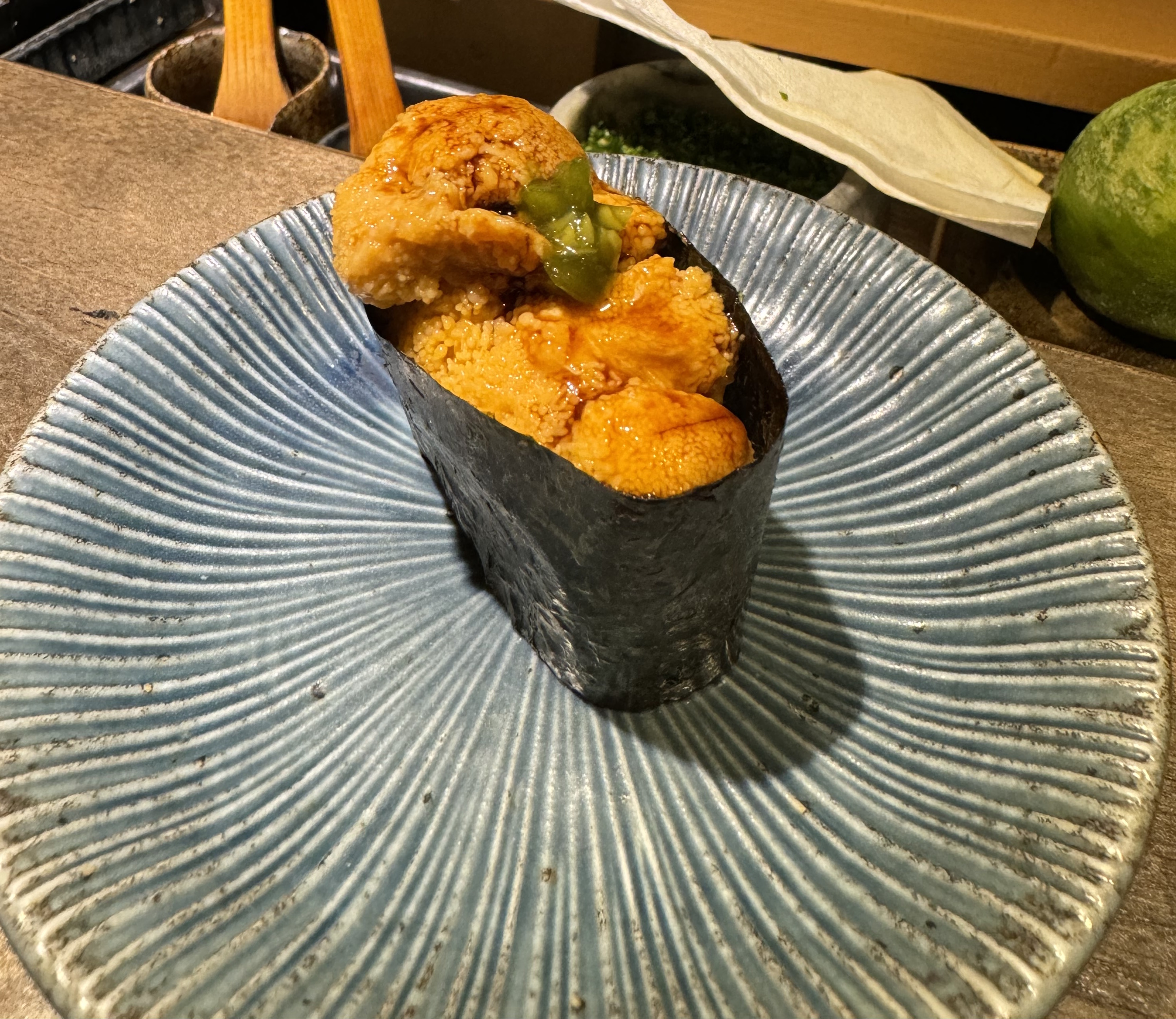 A fairly tall sashimi, with a large piece of nori barding it. The nori barding is to contain the fresh uni: bright yellow tounges, stacked to overflowing, and brushed with a dark brown sauce. There is a green, saucy garnish in the center.