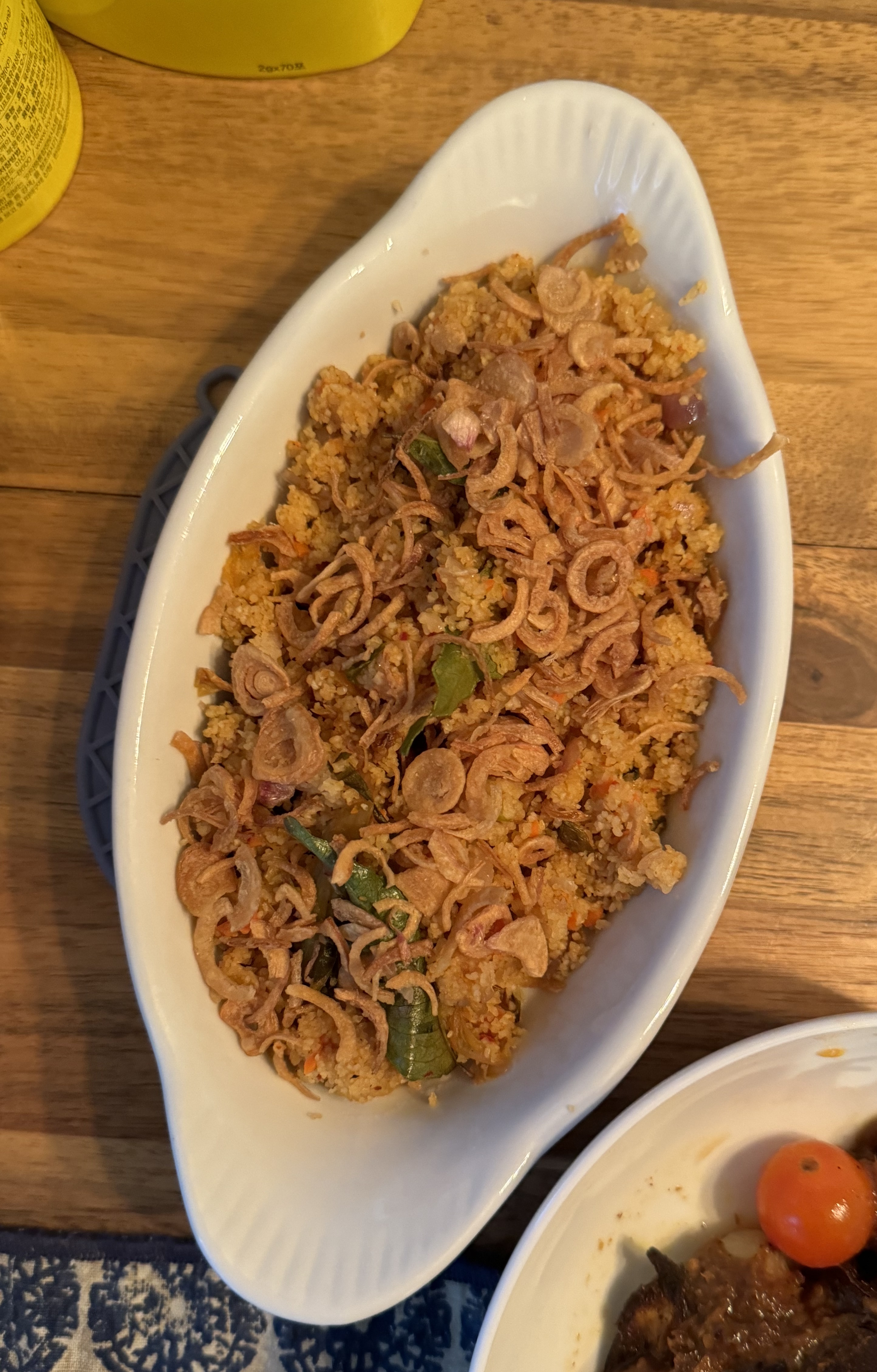 Long dish filled with some fried onions, and other light-brown granules underneath. There are a few curry leaves as garnish. 