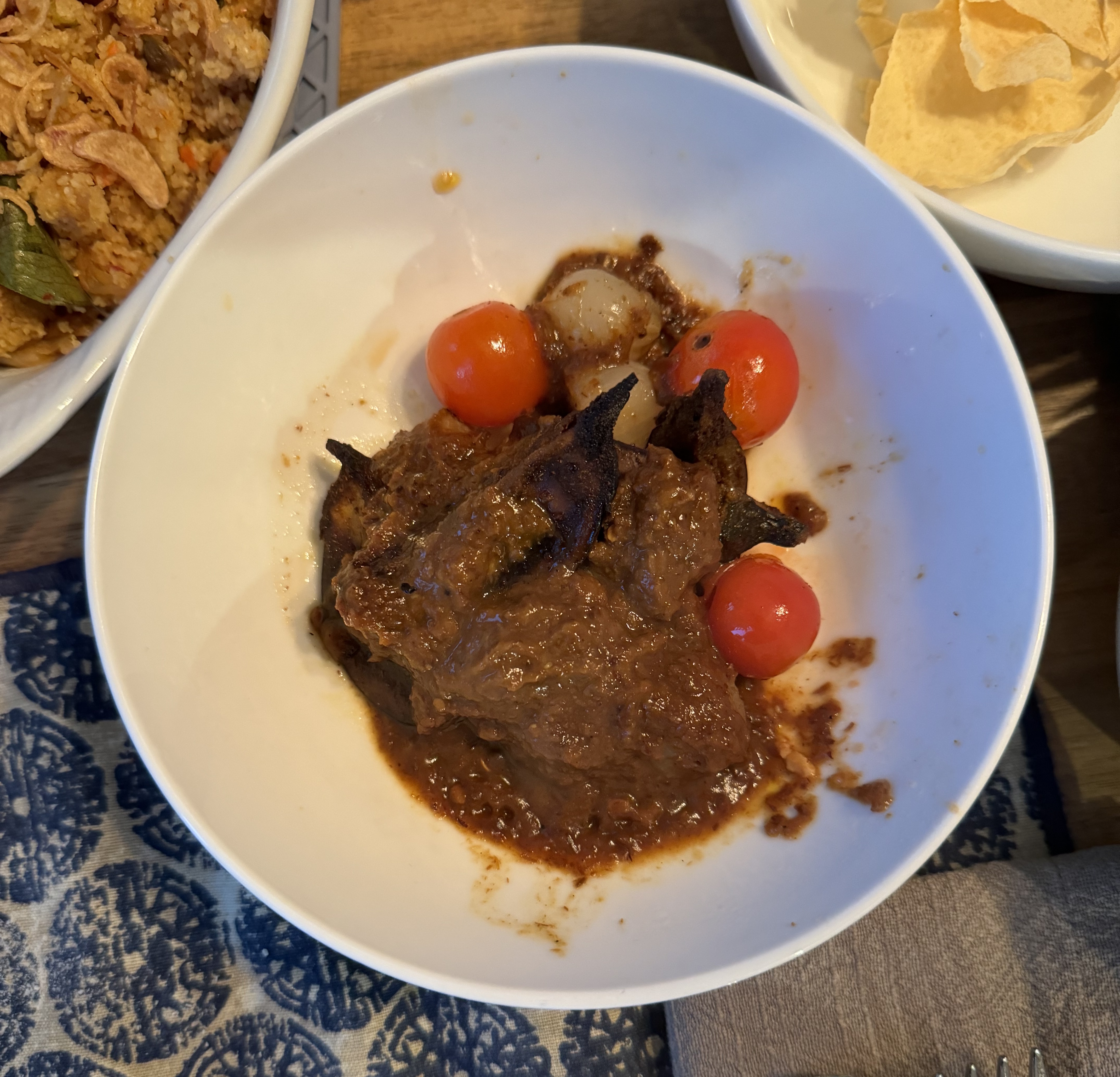 A bowl with some vegetables in a thick brown sauce, which can't really be made out because the sauce is so heavy. There are bright red cherry tomatoes and some cooked pearl onions on the side. Dark tips of curry leaves are poking out of the sauce.