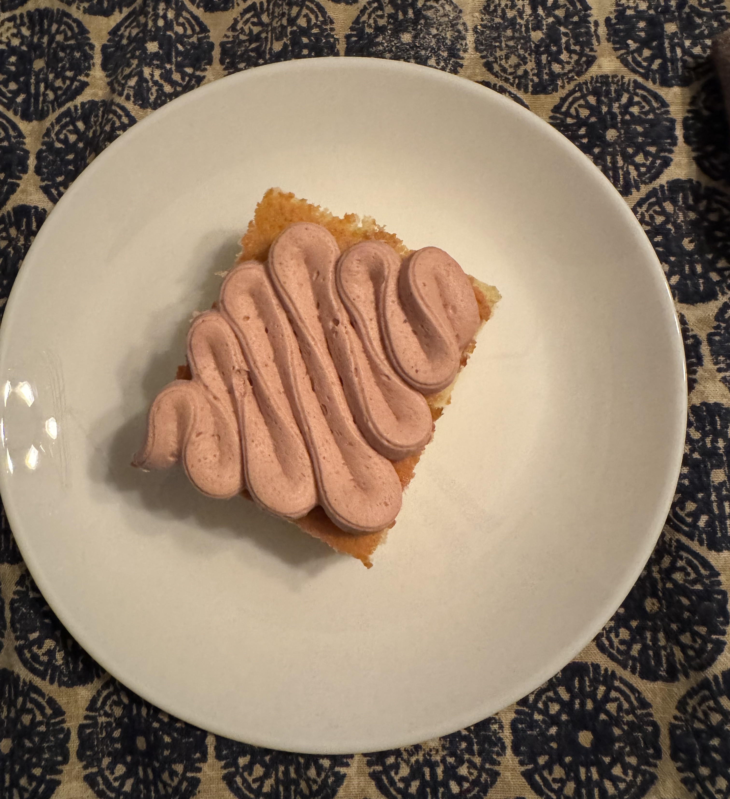 A rectangular slice of yellow cake on a white plate. The frosting is very light pink, and has been piped in a wavy pattern atop the whole slice of cake.