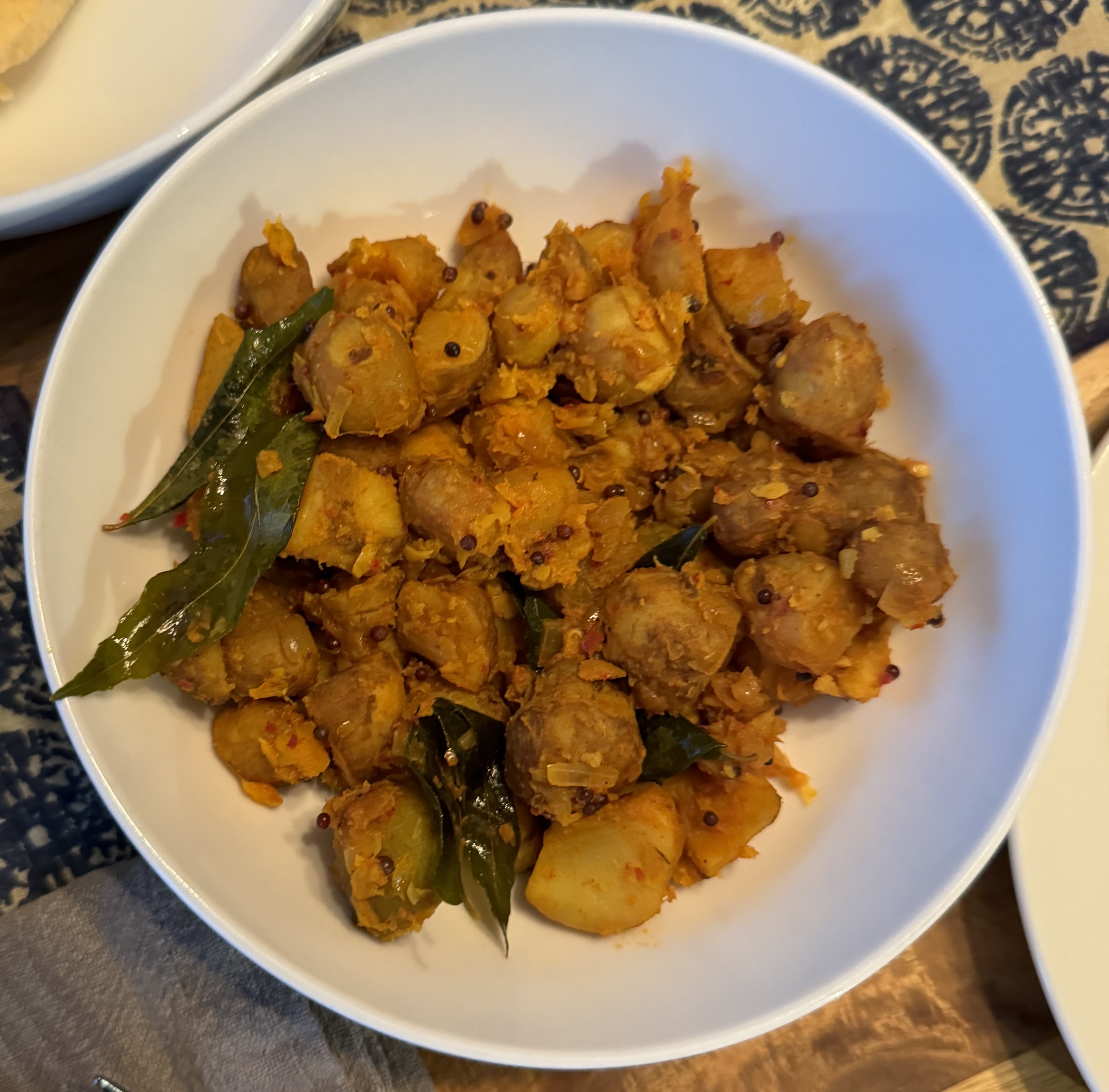 Bowl with small potatoes. The dish is very dry (as opposed to saucy), with some bits of whole spice visible. The potatoes are coated in something opaque, like starch that has been cooked onto them. There are a few curry leaves as garnish.