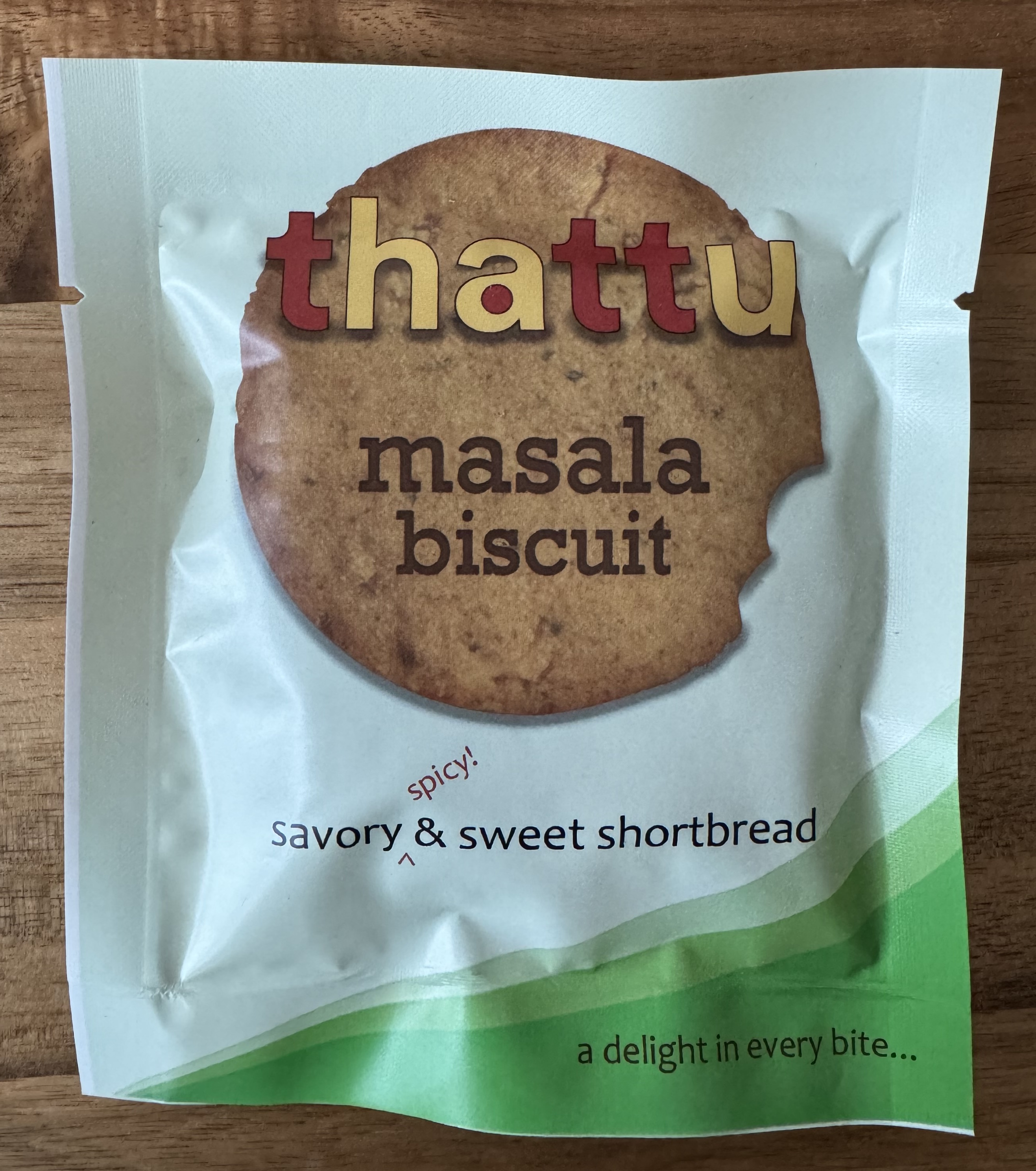 A pre-packed cookie with the Thattu branding. The package has a picture of the cookie inside: large, round, smooth, and brown. It's a masala biscuit: savory, spicy, sweet shortbread.