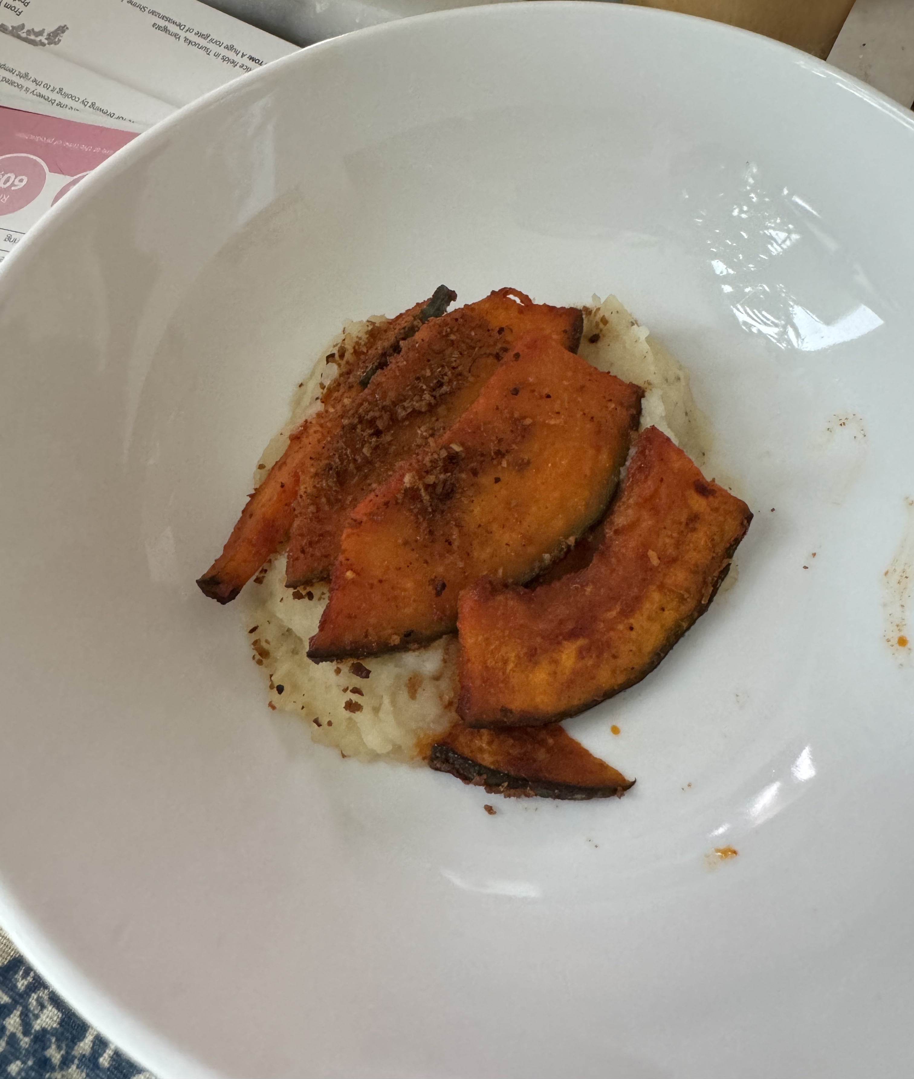 White puree with a few slices of roasted, vibrant orange squash neatly placed on top of it, overlapping each other. The center pieces of squash have a crumble on them.