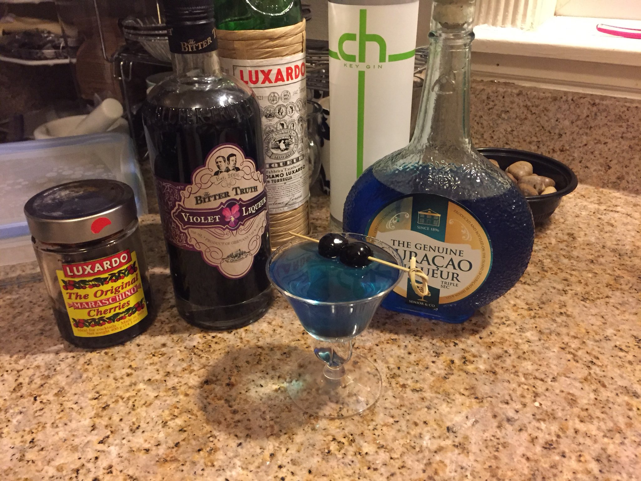 Blue cocktail in a small nick-and-nora style couple glass, with two dark cherries on top.

A bottle of violet liquor and curacao are behind them. There is a jar of cocktail cherries.