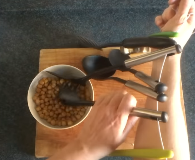 A man's arm with a cord tied from shoulder to hand. Various kitchen implements, like large spoons and mashers, are hung on the string. He is using one very awkwardly to wangjangle a bowl of chickpeas.